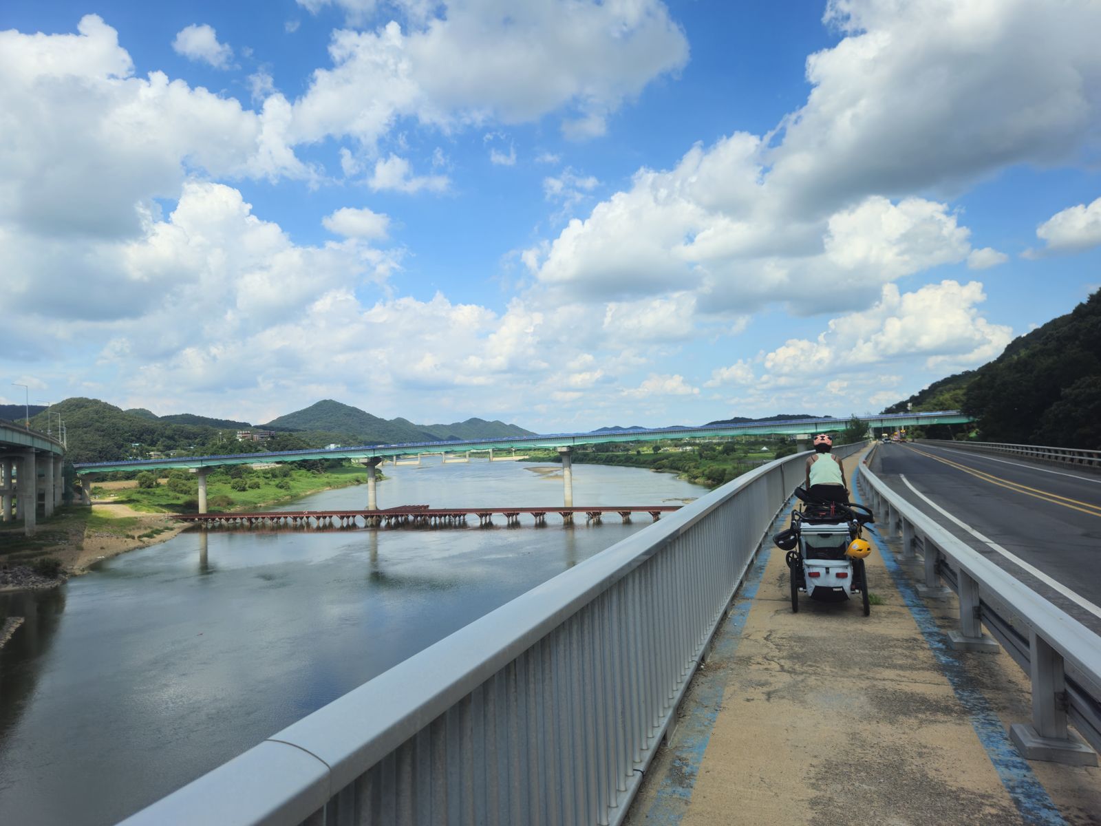 Un cycliste avec une remorque traverse un pont sur la rivière Geum, entouré de collines verdoyantes et d'un ciel nuageux.