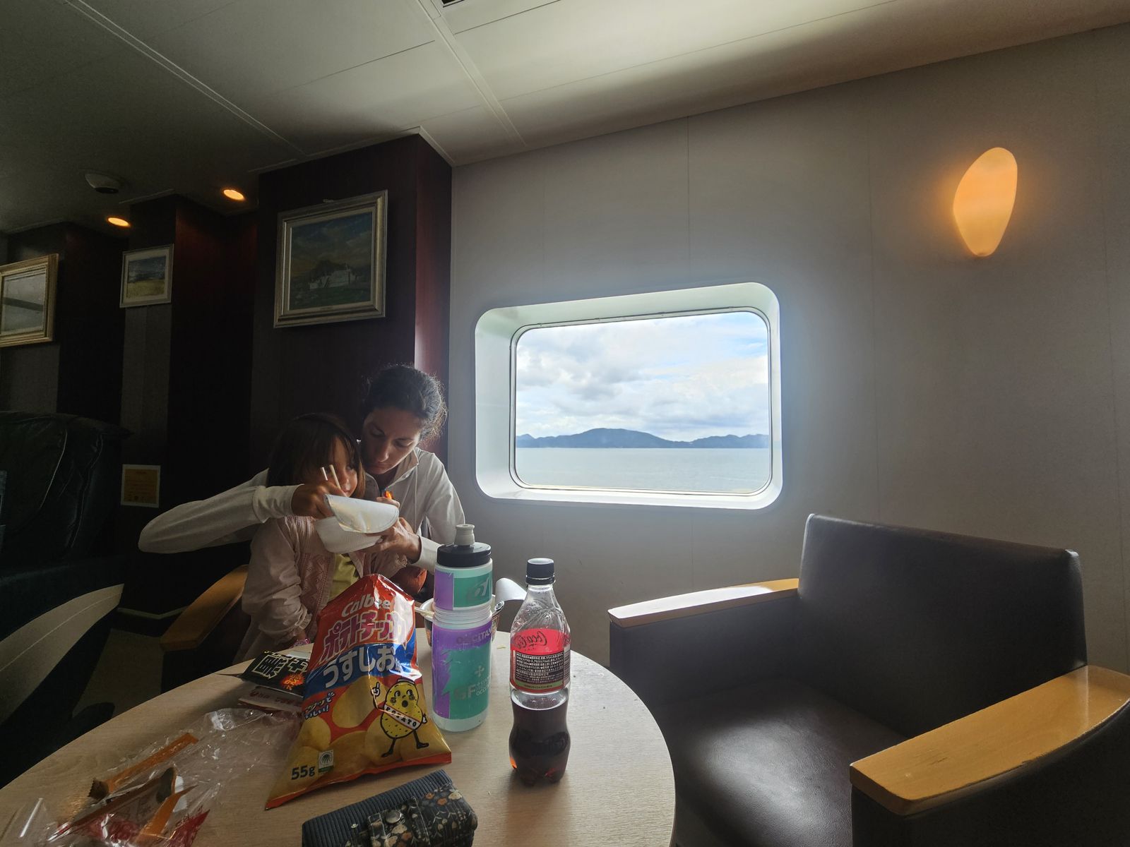Une mère et sa fille partagent un repas à bord d'un ferry, avec une vue sur la mer à travers une grande fenêtre.