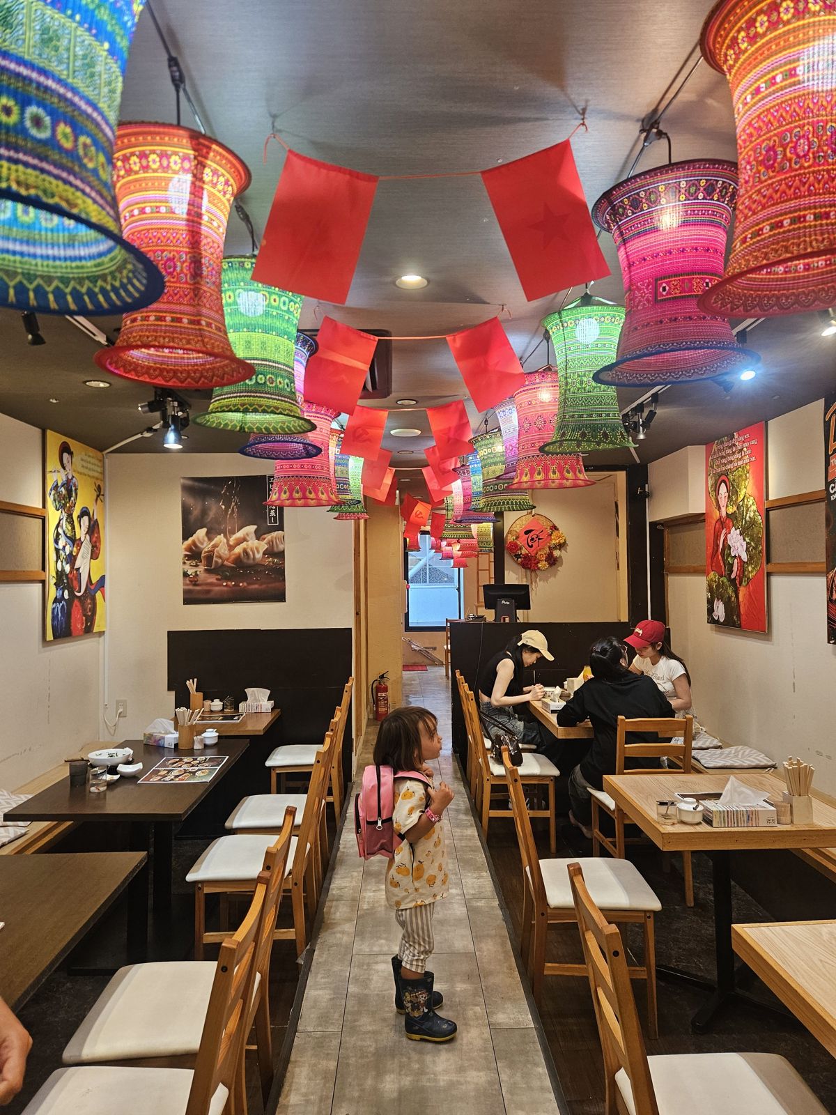 Intérieur coloré d'un restaurant avec des lanternes décoratives et des drapeaux. Une petite fille avec un sac à dos se tient debout au centre, regardant autour d'elle.