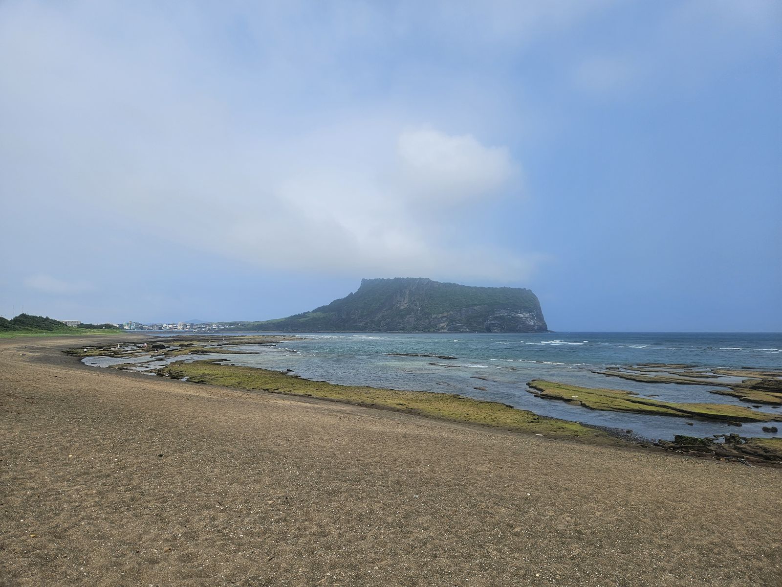 Vue panoramique de la plage avec des formations rocheuses et une colline à l'horizon, sous un ciel nuageux.