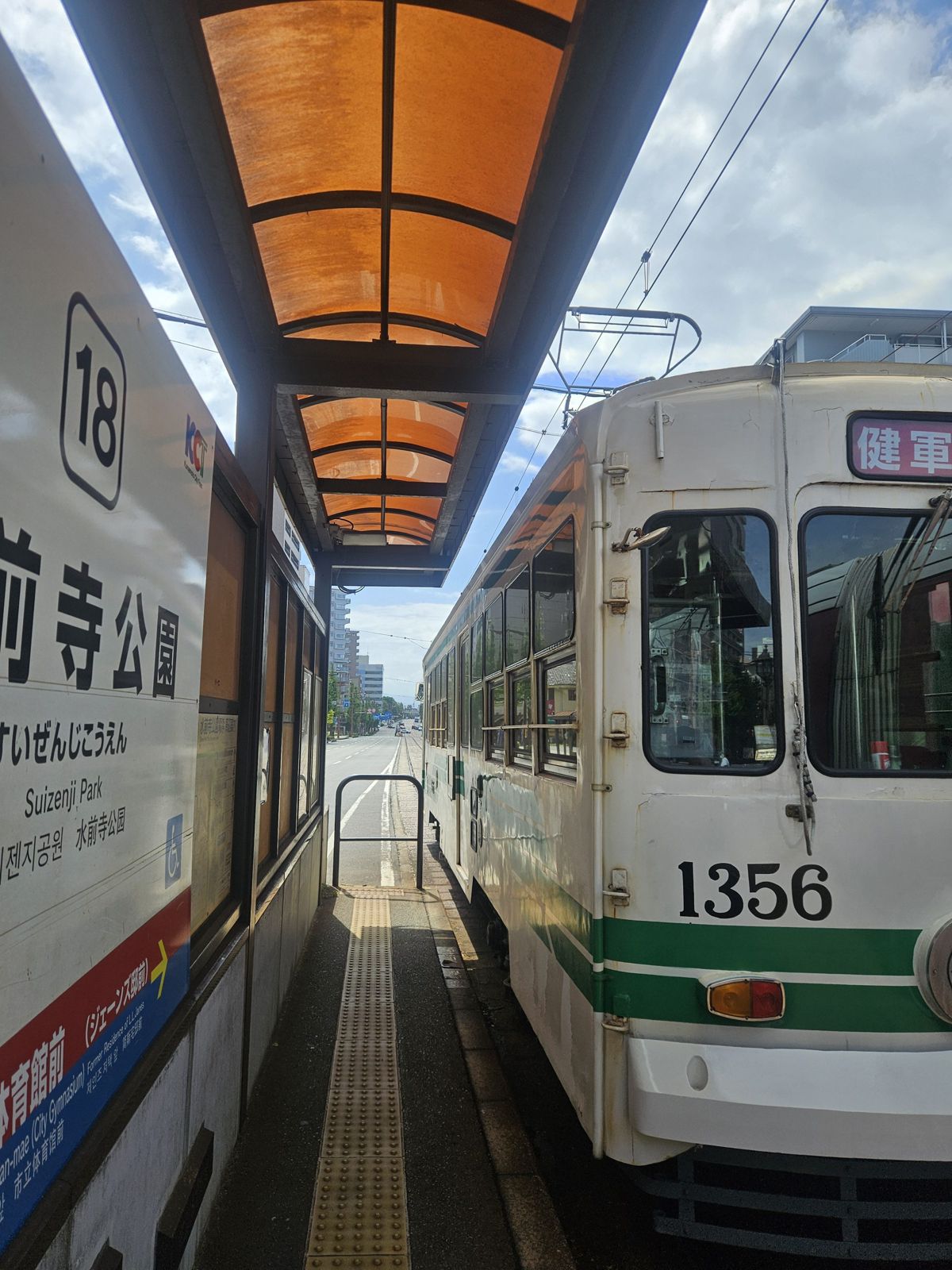 Un tramway à l'arrêt à la station Suizenji Park, avec un toit orange, sous un ciel partiellement nuageux.