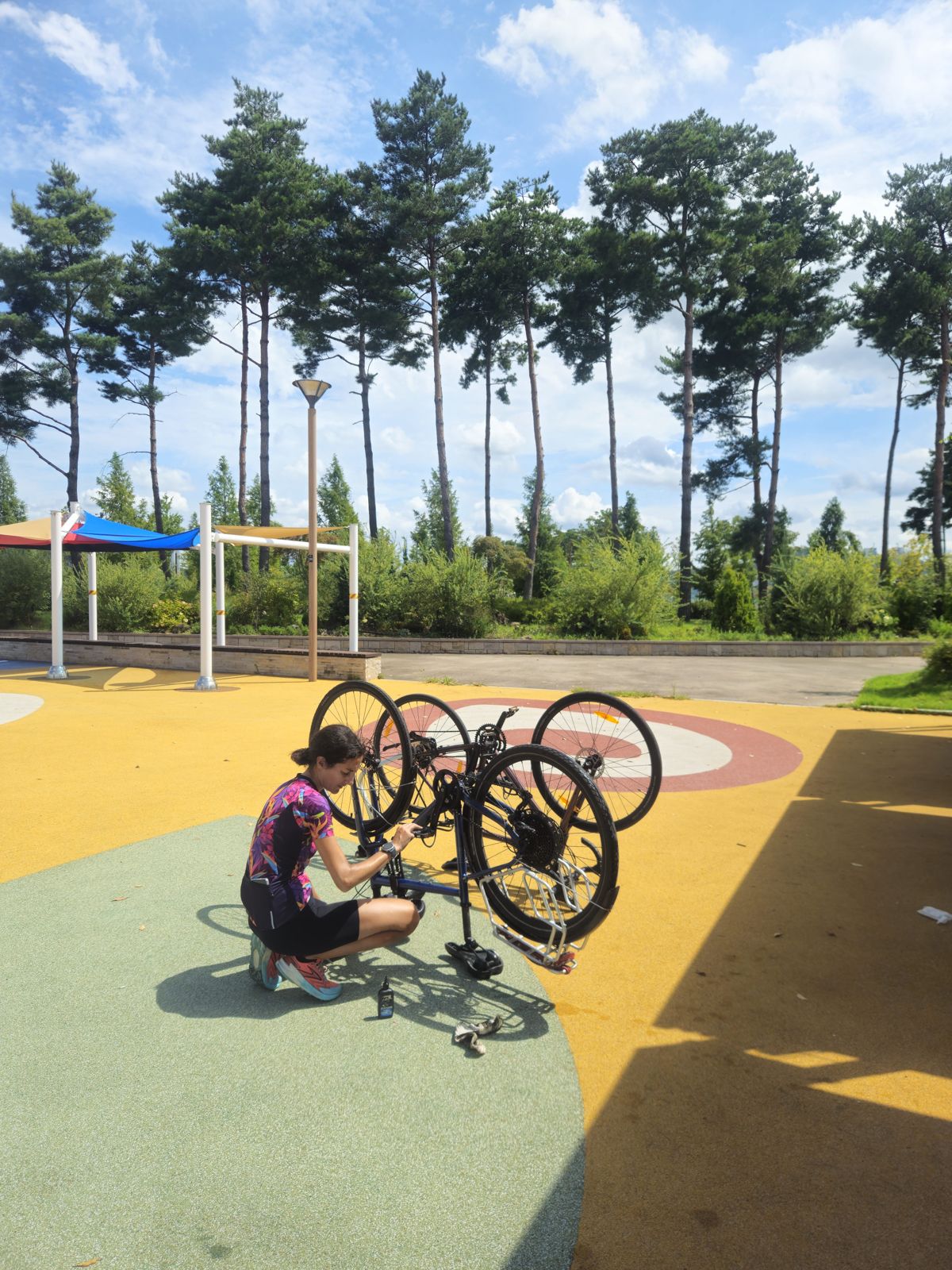 Une enfant répare un vélo sur un terrain de jeu en plein air, entourée de grands arbres et d'un ciel bleu.