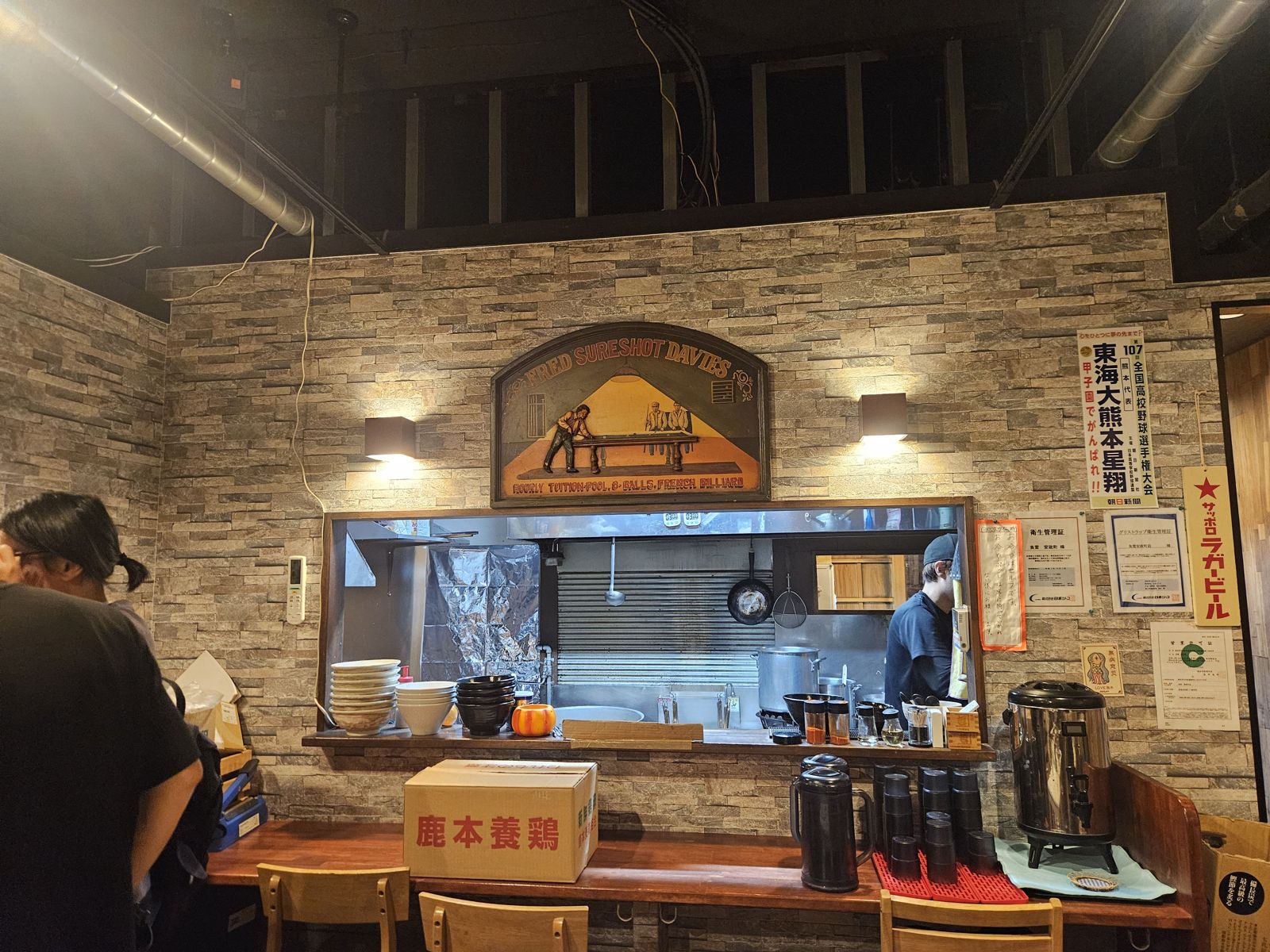Intérieur d'un restaurant avec des murs en pierre, affiches et comptoir où un chef prépare des plats. Des assiettes et tasses sont visibles sur la table.
