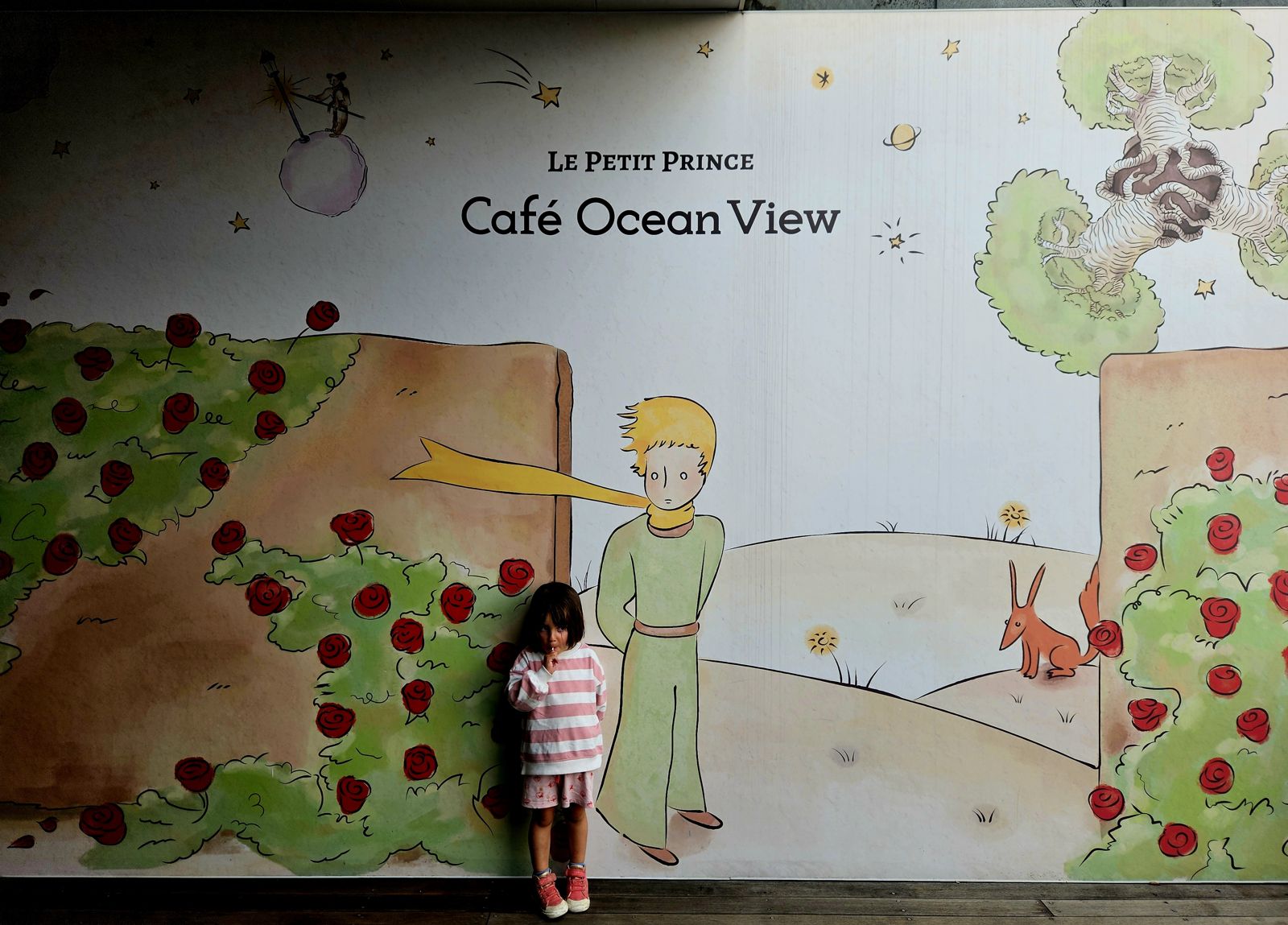 Une petite fille se tient devant une murale colorée représentant Le Petit Prince, avec des roses et un renard, dans un café nommé 'Café Ocean View'.