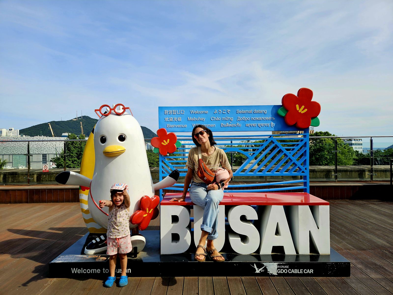 Une petite fille souriante posant avec une mascotte en forme de pingouin, devant un panneau 'Welcome to Busan', avec des fleurs colorées en arrière-plan.