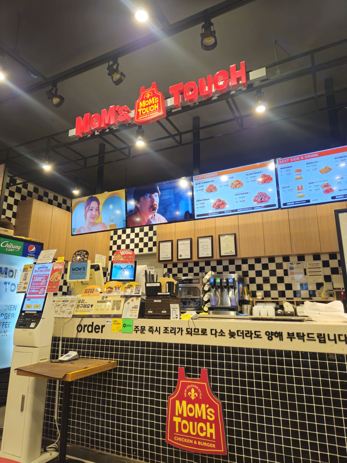 Intérieur d'un restaurant de fast-food Mom's Touch, avec un comptoir de commande et des écrans affichant le menu.