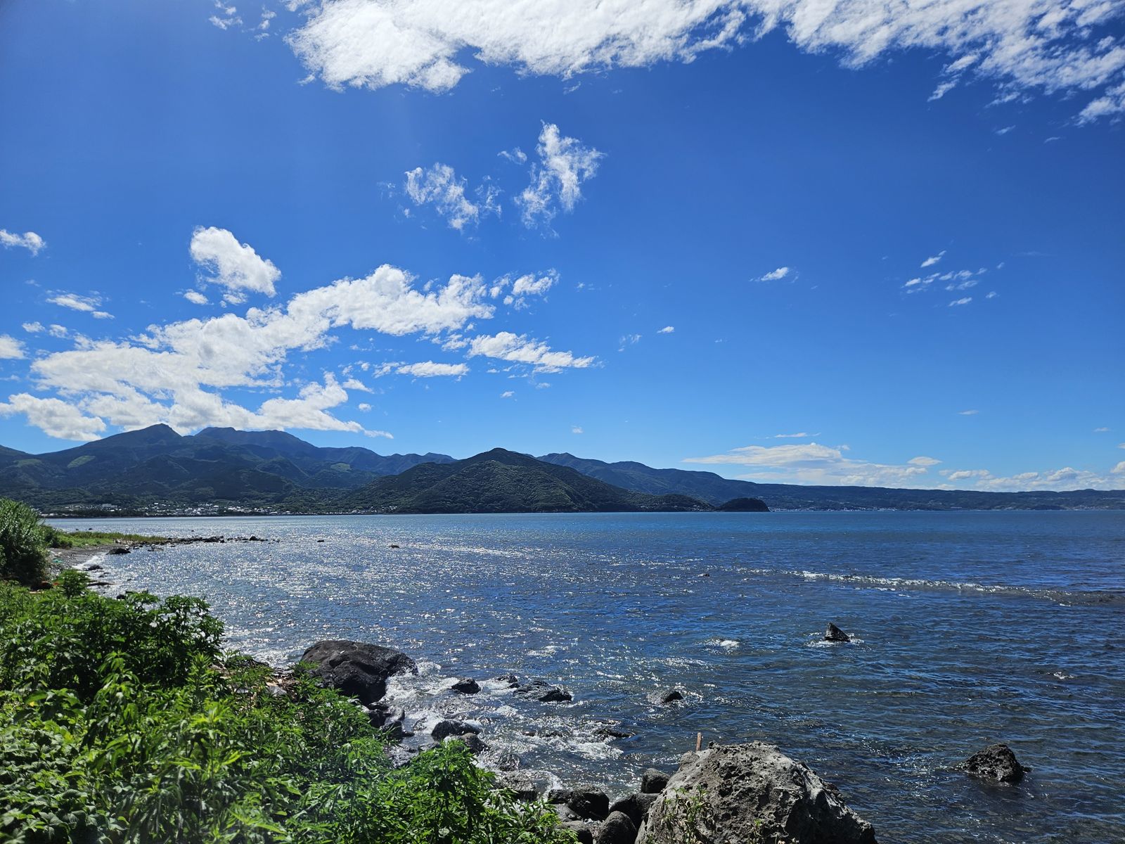 Vue panoramique sur la mer avec des montagnes en arrière-plan sous un ciel bleu parsemé de nuages blancs.