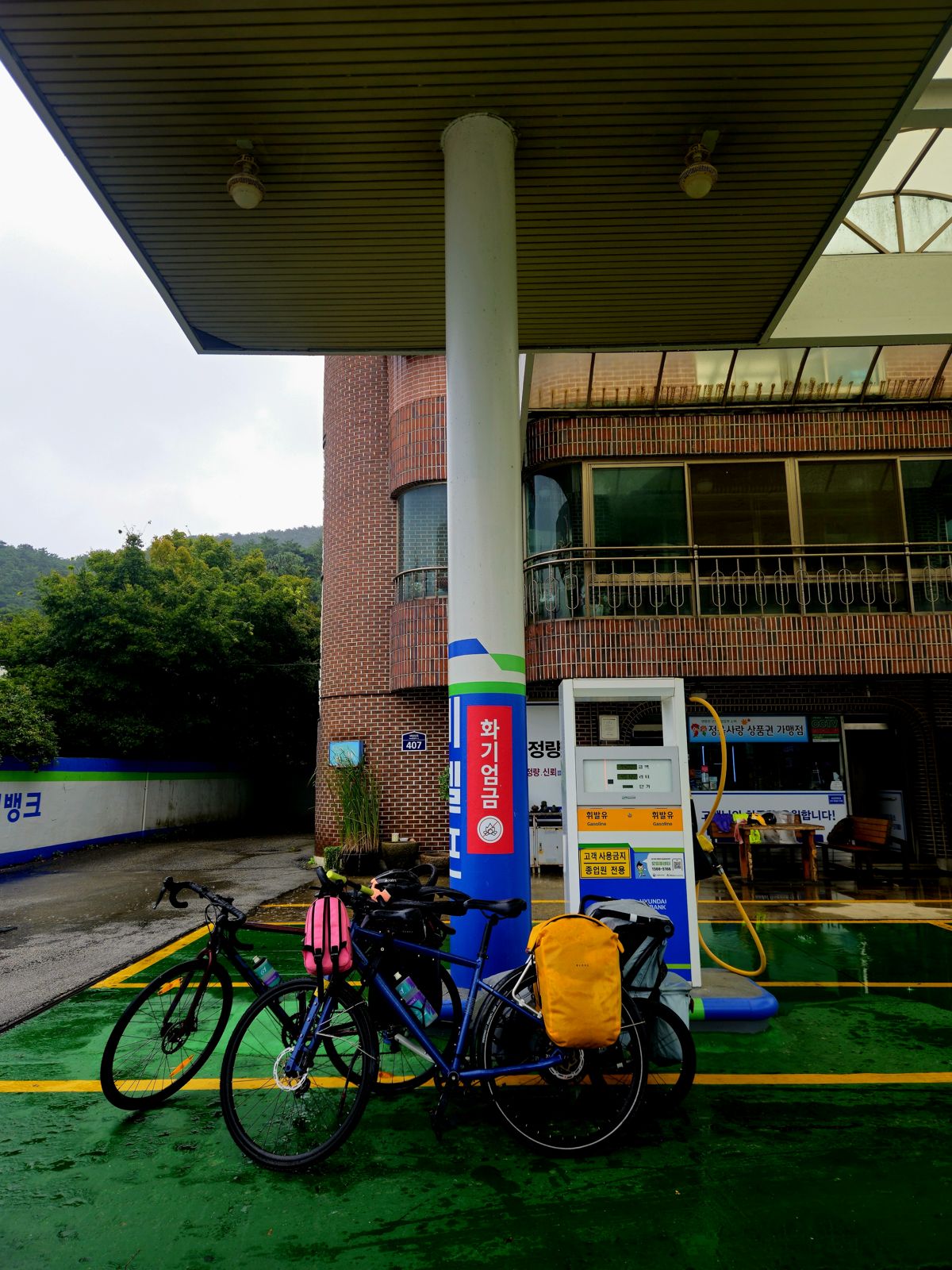 Deux vélos avec des sacoches colorées garés sous un abri près d'une station-service sous la pluie.