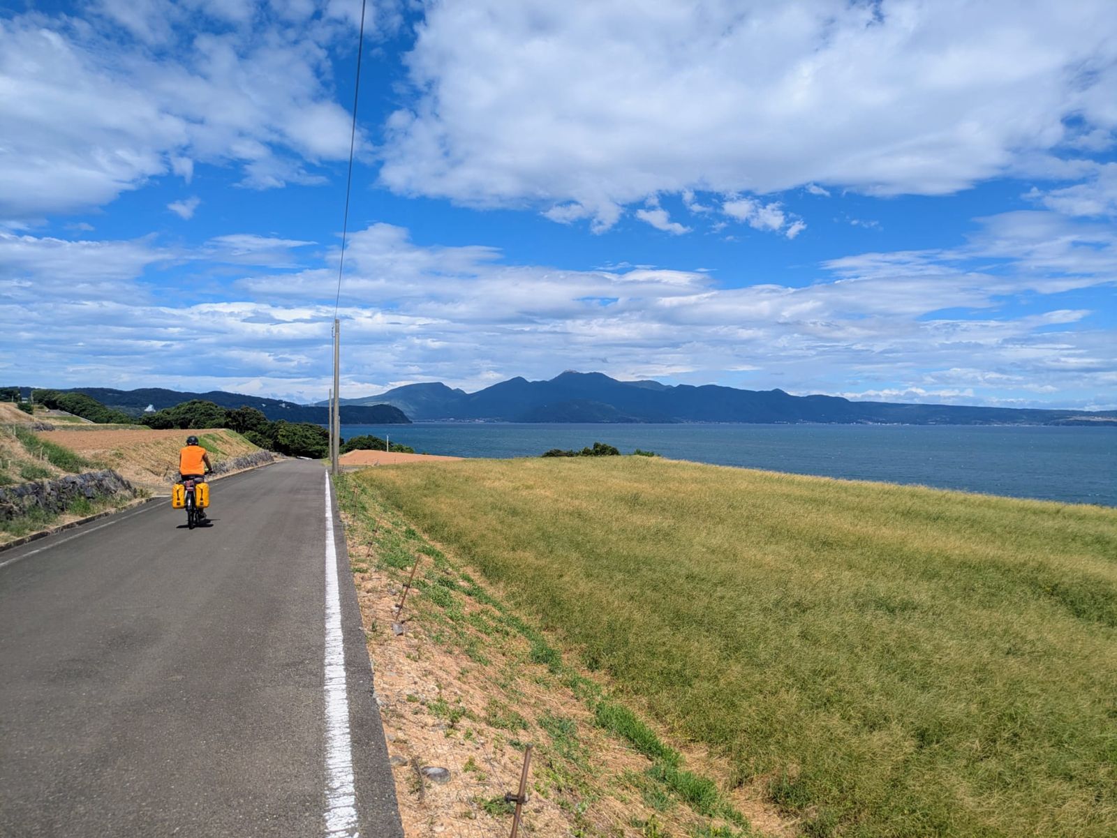 Un cycliste sur une route bordée de champs et surplombant la mer, avec des collines en arrière-plan sous un ciel partiellement nuageux.