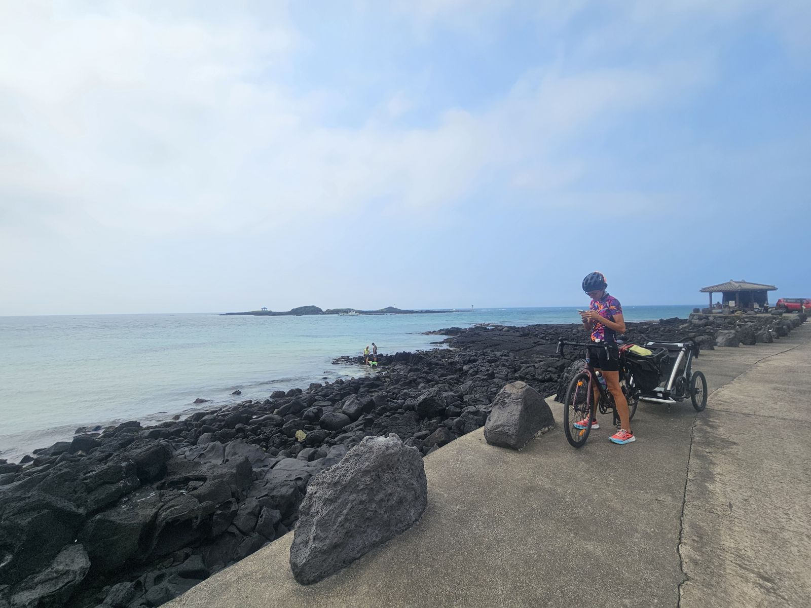 Femme en tenue de vélo regardant son téléphone sur un chemin en bord de mer, avec des rochers noirs et une eau turquoise en arrière-plan.