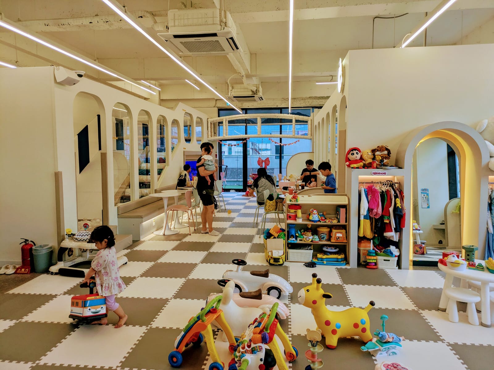 Intérieur d'un café pour enfants, avec des enfants jouant et des parents présents. Le sol est en dalles de mousse gris et blanc, et plusieurs jouets colorés sont éparpillés.