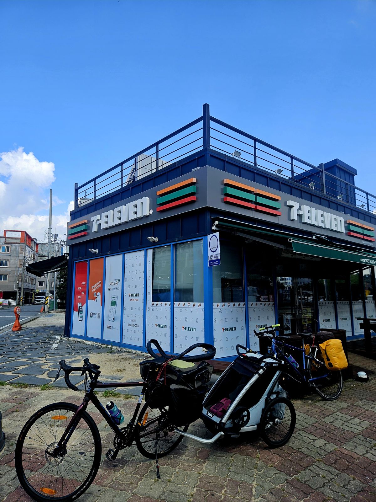 Vue d'un magasin 7-Eleven avec un ciel bleu en arrière-plan, accompagné de plusieurs vélos garés devant.