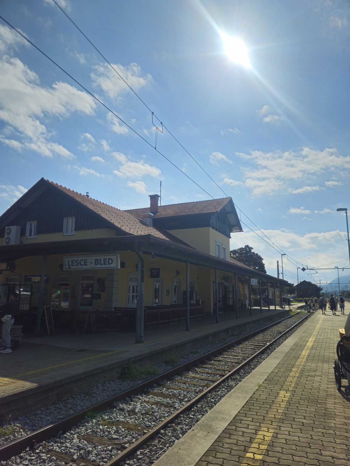 Gare de Lesce-Bled sous un ciel ensoleillé avec des nuages, affichant une architecture traditionnelle et des rails de train.