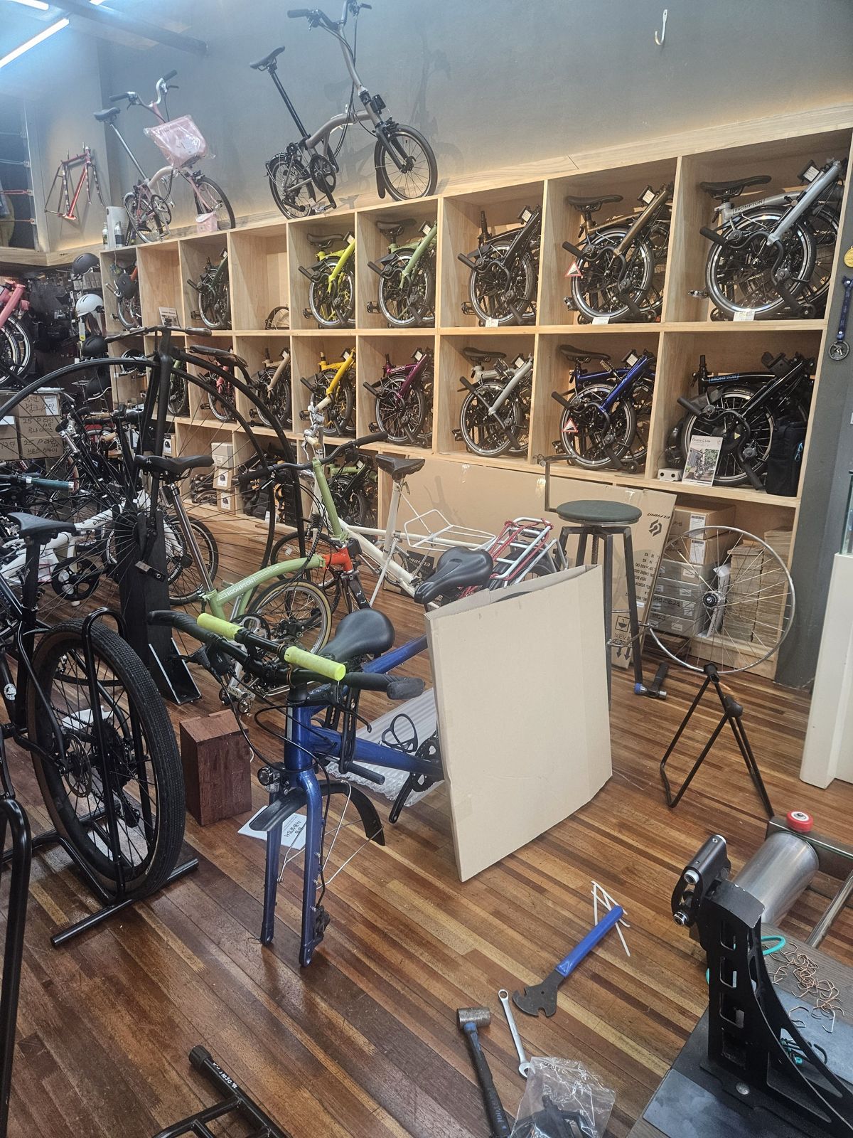 Intérieur d'un magasin de vélos avec des vélos pliants exposés sur des étagères en bois. Le sol en bois est encombré d'outils et de pièces de vélo.