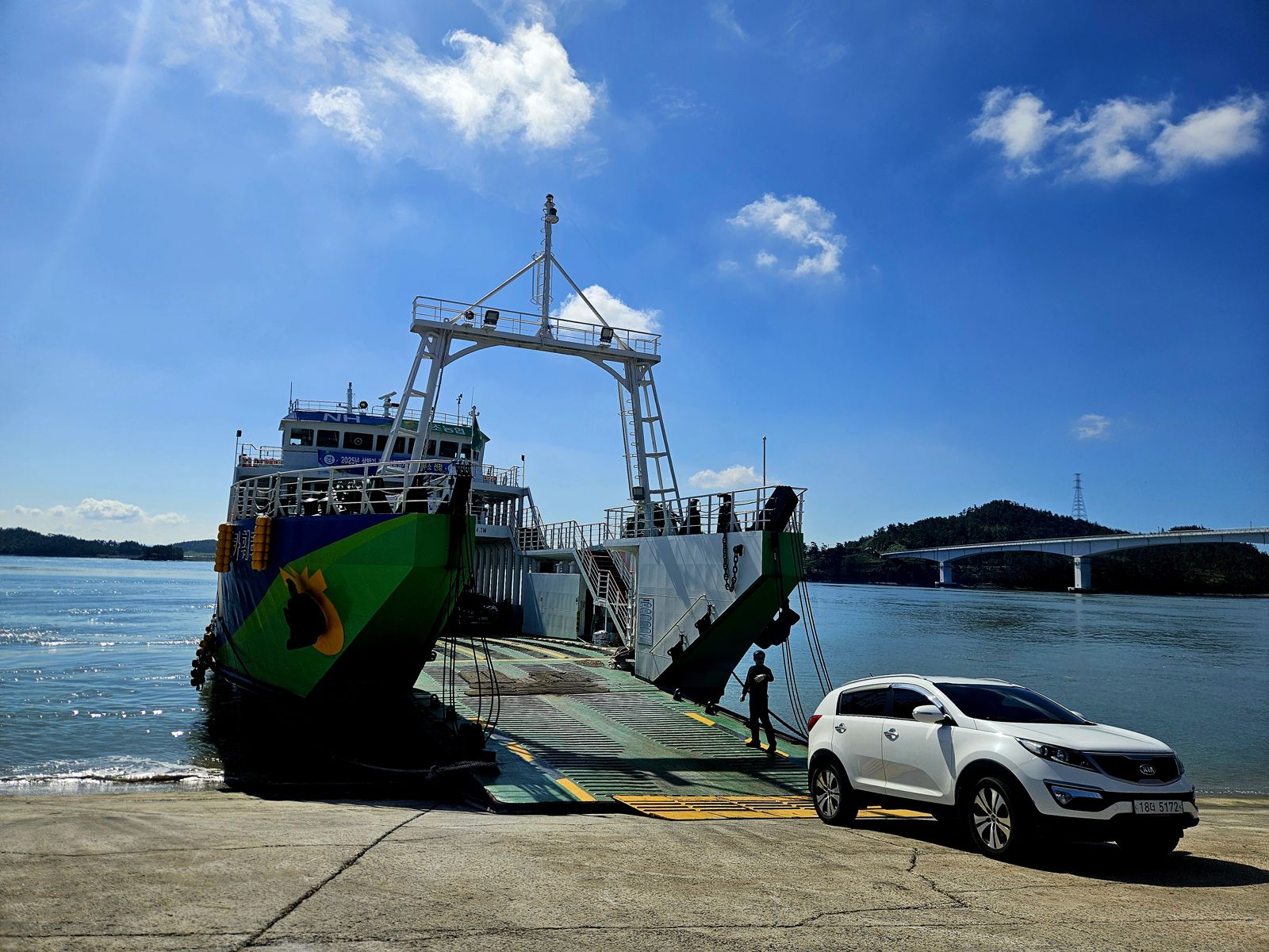 Un ferry coloré à quai dans un terminal presque vide, avec une voiture blanche stationnée à proximité et un ciel bleu en arrière-plan.