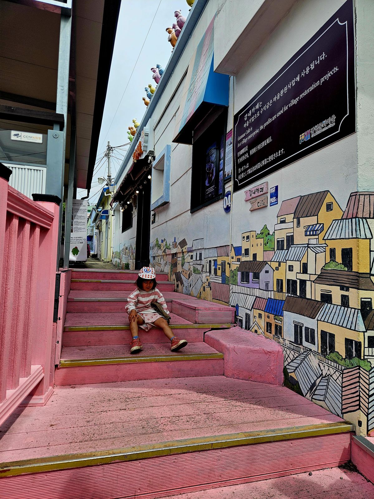 Une enfant assise sur des marches colorées, entourée de murs peints représentant des maisons. Le décor est vibrant avec des couleurs vives et une ambiance artistique, illustrant un ancien village portuaire rénové.