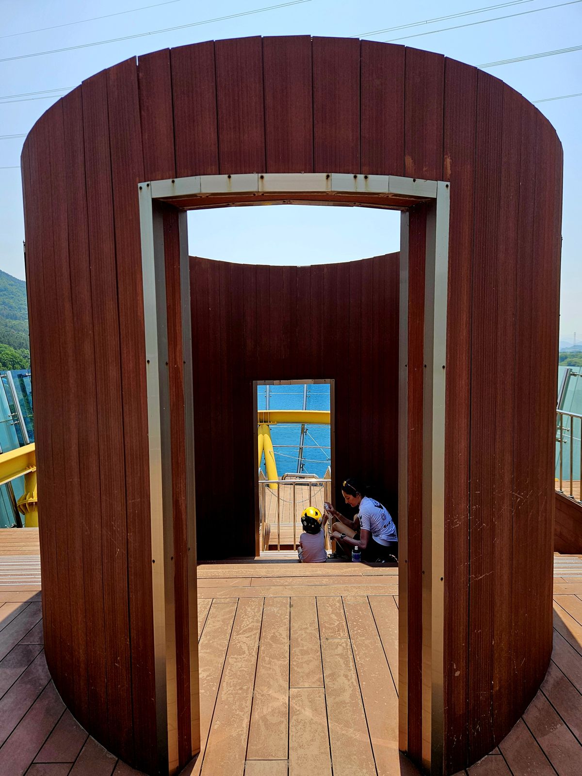 Une mère et son enfant assis dans une structure architecturale en bois, avec une vue sur un plan d'eau au fond, par une journée ensoleillée.