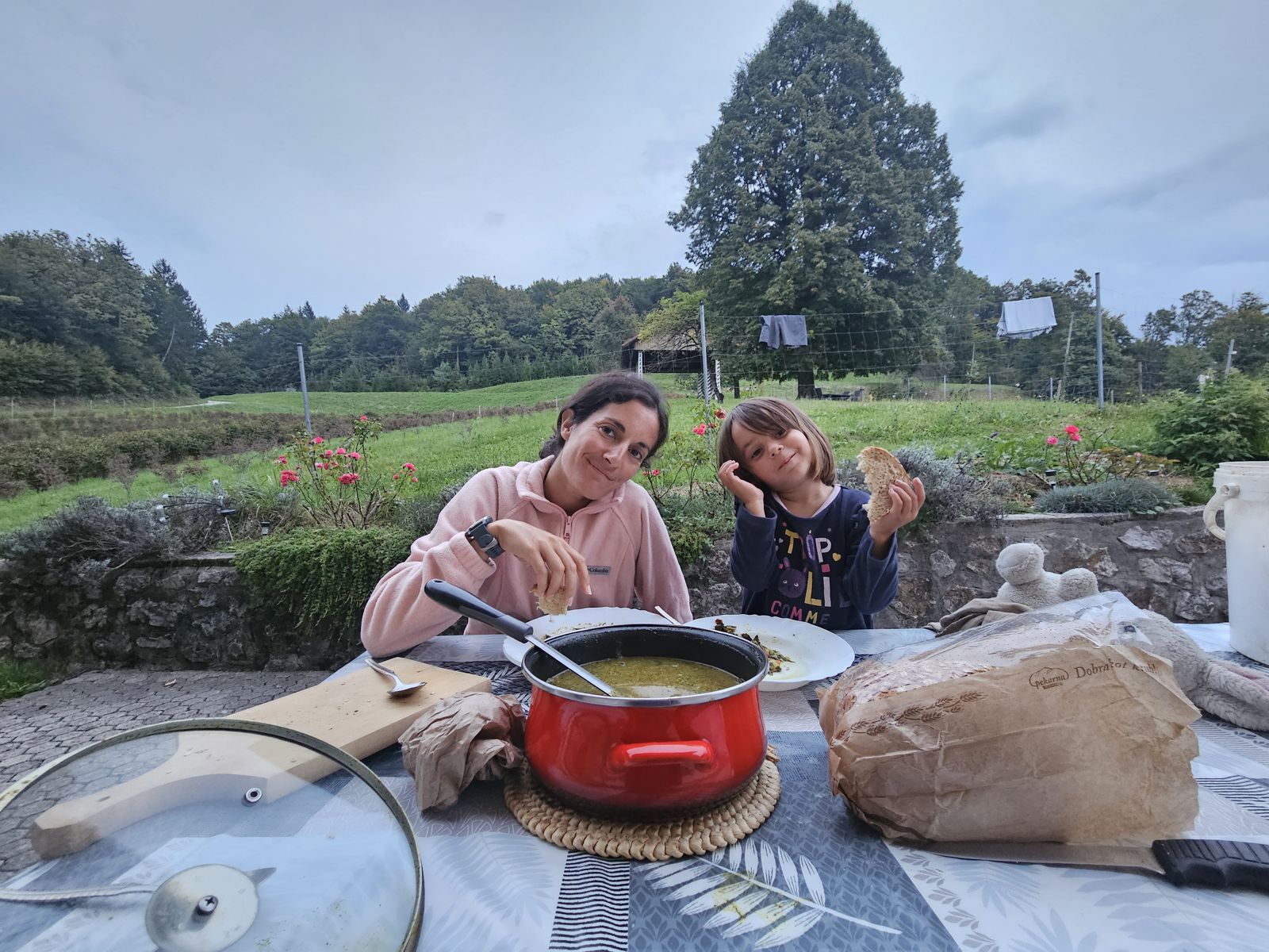 Deux personnes, une femme et une petite fille, sourient à la caméra en prenant leur repas en plein air, avec un plat en pot rouge au centre de la table et des fleurs autour.