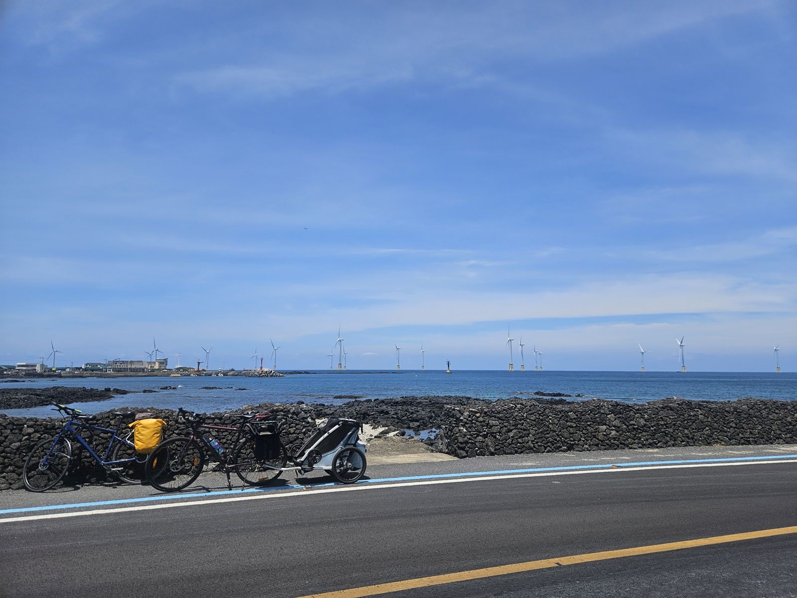 Un paysage côtier de Jeju, Corée du Sud, avec des vélos garés sur le bord de la route. Des éoliennes sont visibles au loin sur la mer. Le ciel est dégagé et bleu.