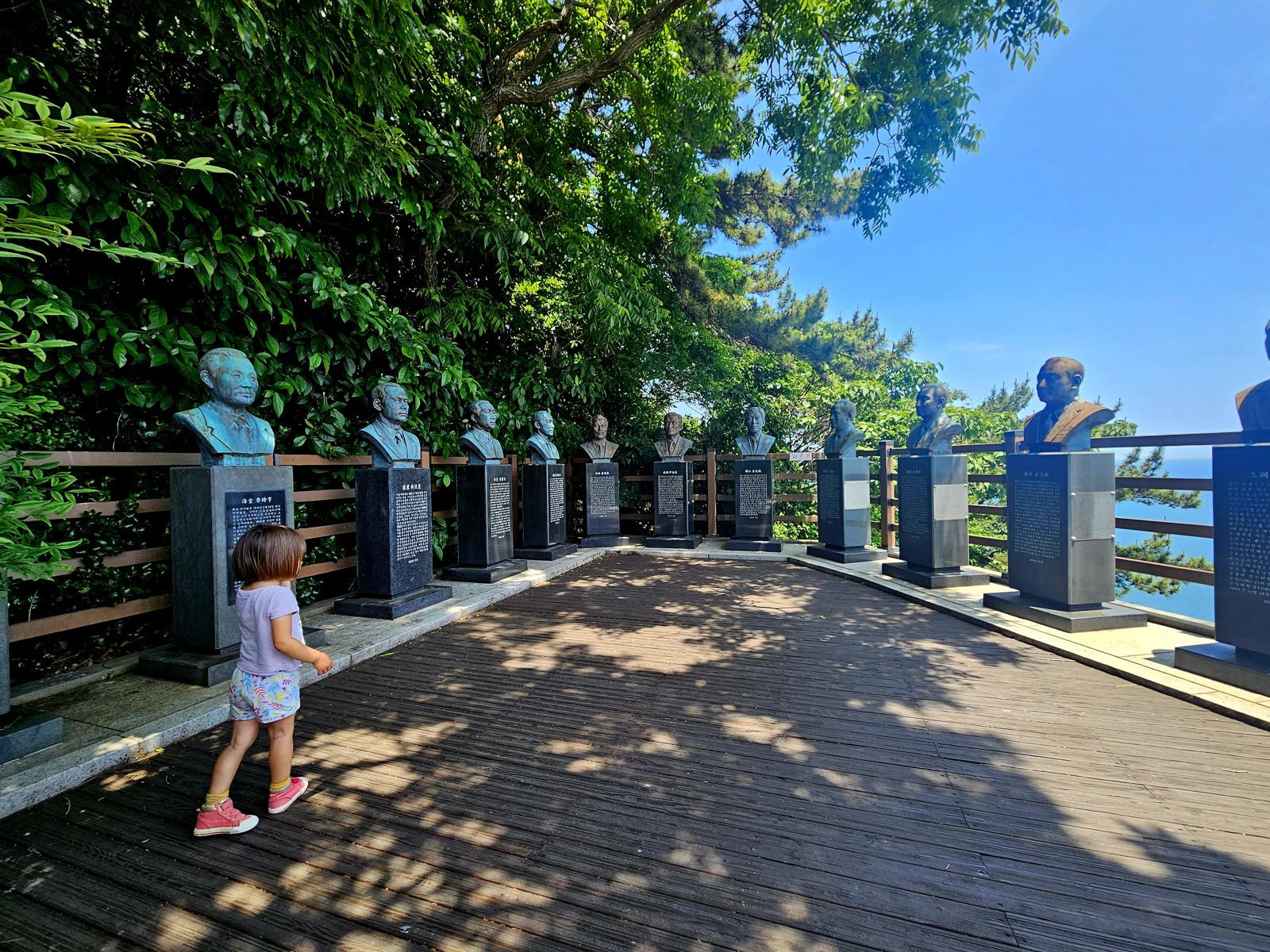Une petite fille marche sur une plateforme en bois, entourée de bustes monumentaux de personnalités. L'environnement est verdoyant avec des arbres en arrière-plan et un ciel bleu.