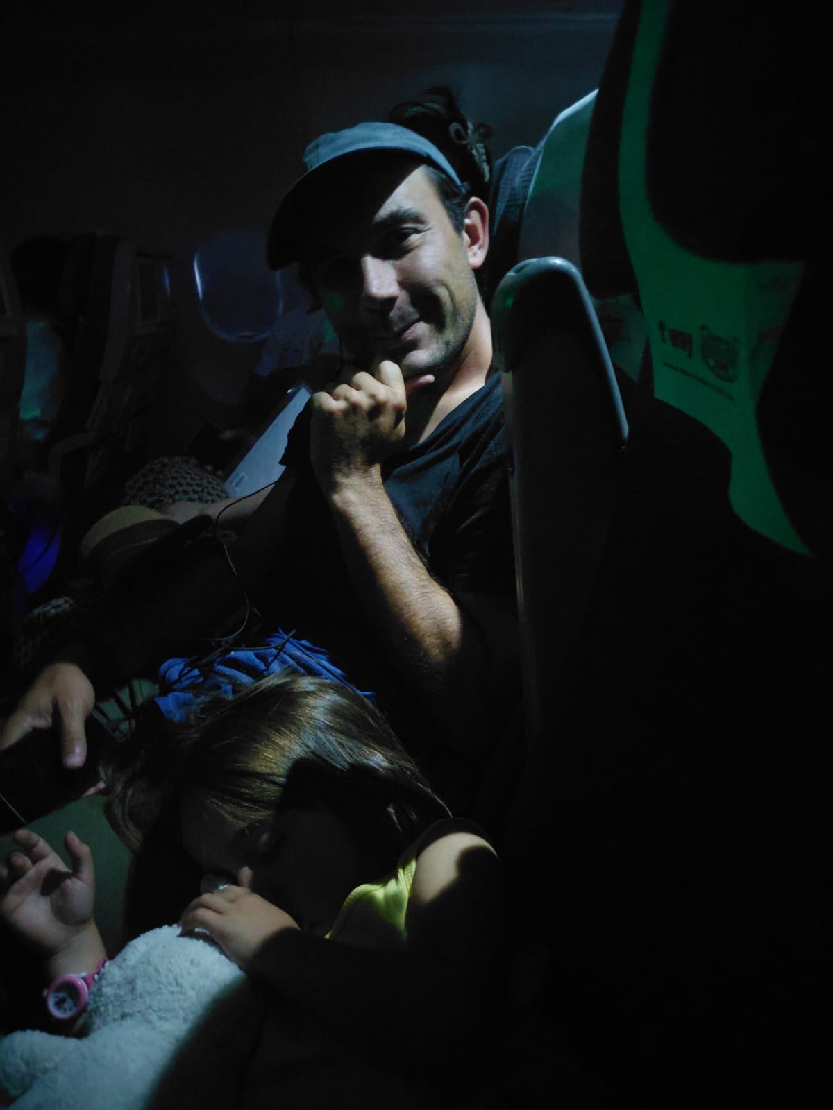 Un homme souriant, assis dans un avion, tenant un écouteur, avec une jeune fille endormie sur son épaule.