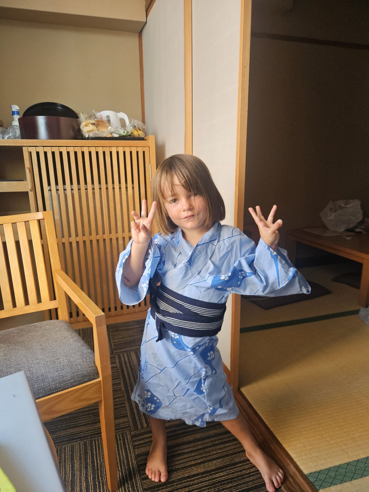 Une petite fille en kimono bleu avec des motifs japonais, se tenant dans une chambre d'hôtel, montrant des signes de paix avec ses mains.