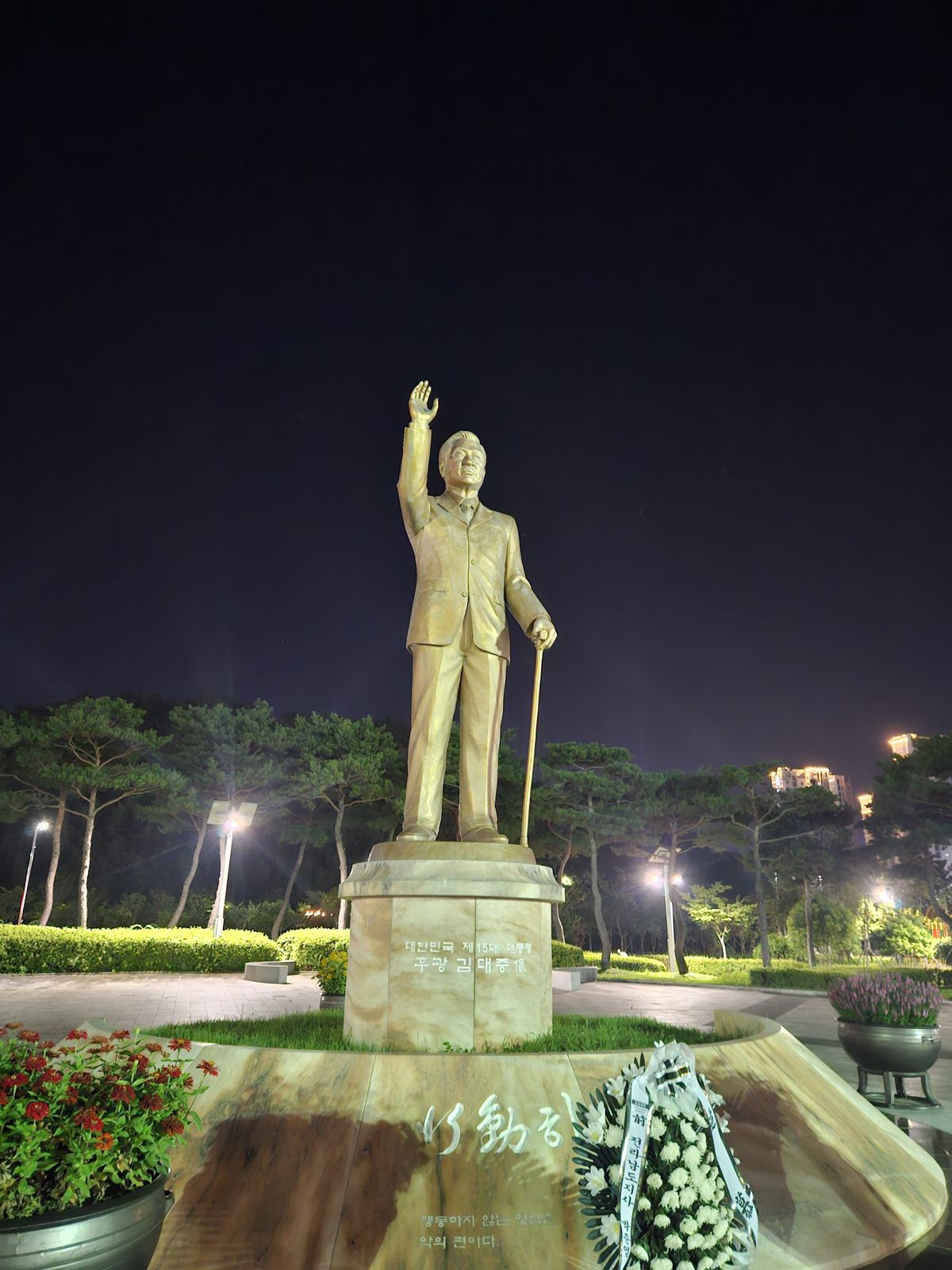 Statue d'une figure historique levant la main, éclairée par des lumières de parc la nuit, entourée de verdure et de fleurs.