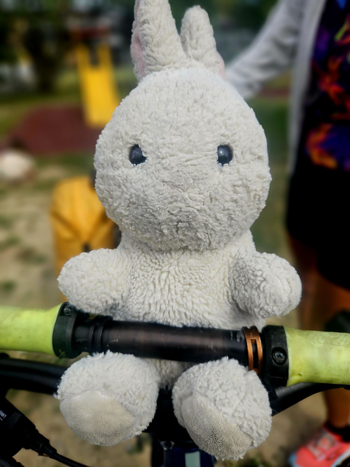 Un jouet en peluche de lapin blanc assis sur le guidon d'un vélo, avec un arrière-plan flou d'une aire de jeux.