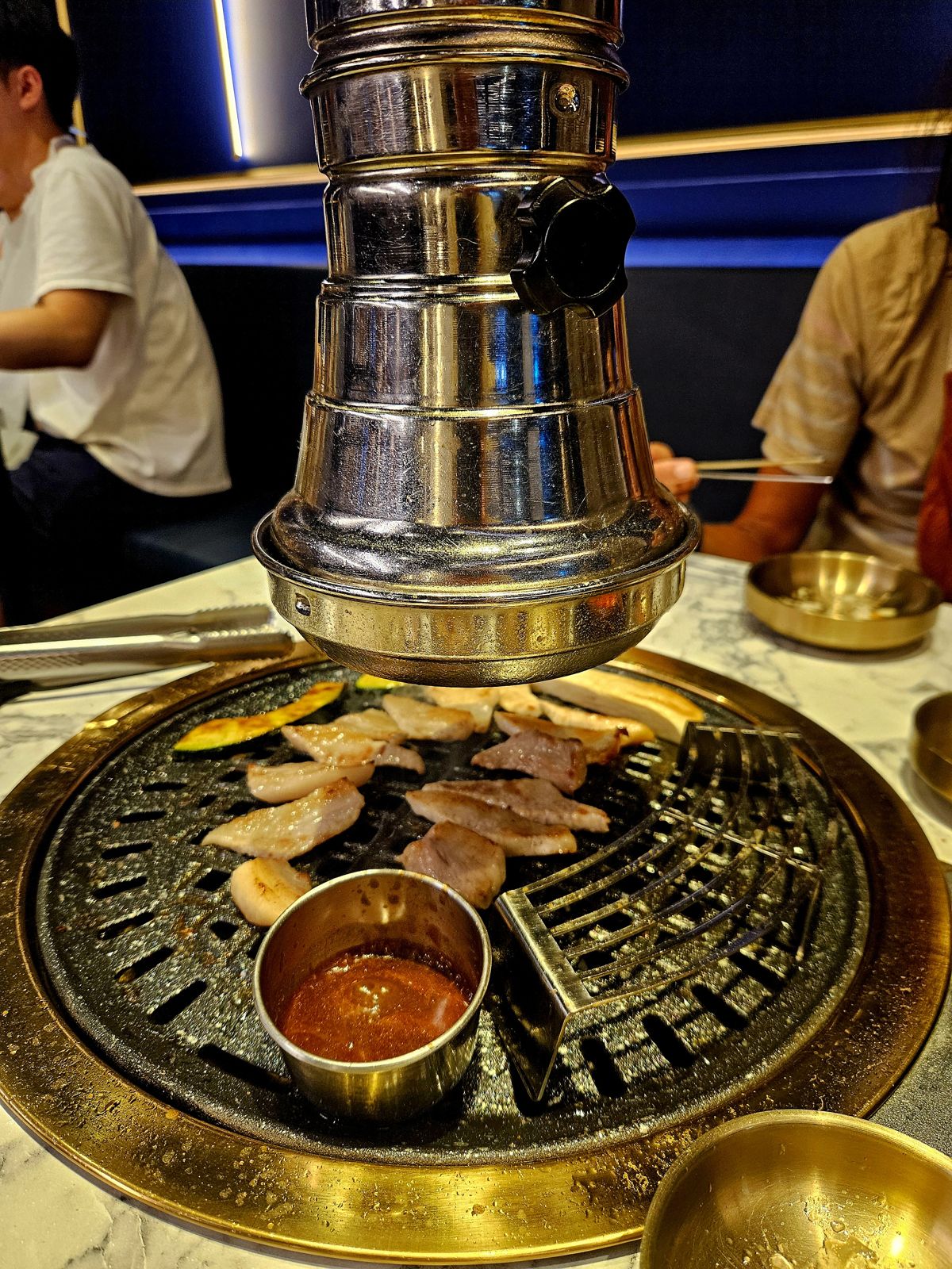 Grill de barbecue coréen avec viande cuisant sur une plaque, sauce dans un bol en métal au premier plan, ambiance de restaurant visible en arrière-plan.