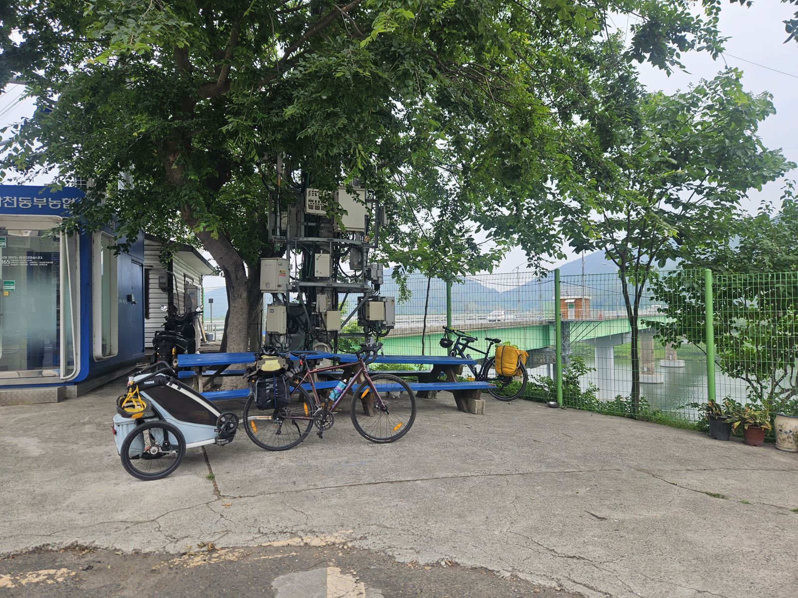 Un espace de repos le long d'une route, avec plusieurs vélos stationnés, dont un avec une remorque. En arrière-plan, un pont vert et des montagnes au loin, sous un ciel nuageux.