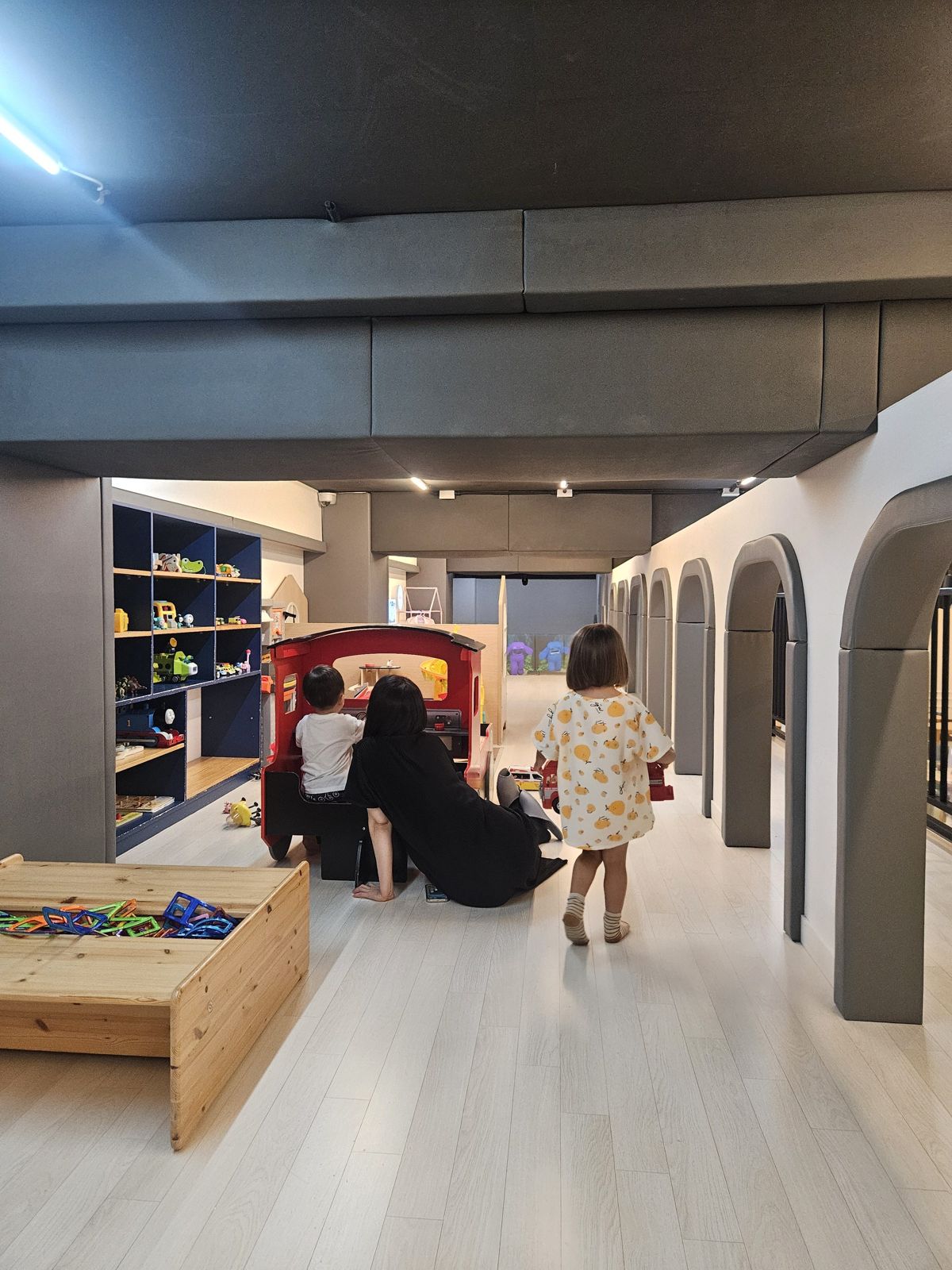 Un espace de jeu intérieur pour enfants avec des jouets colorés, deux enfants jouant dans une voiture, et une adulte assise sur le sol.