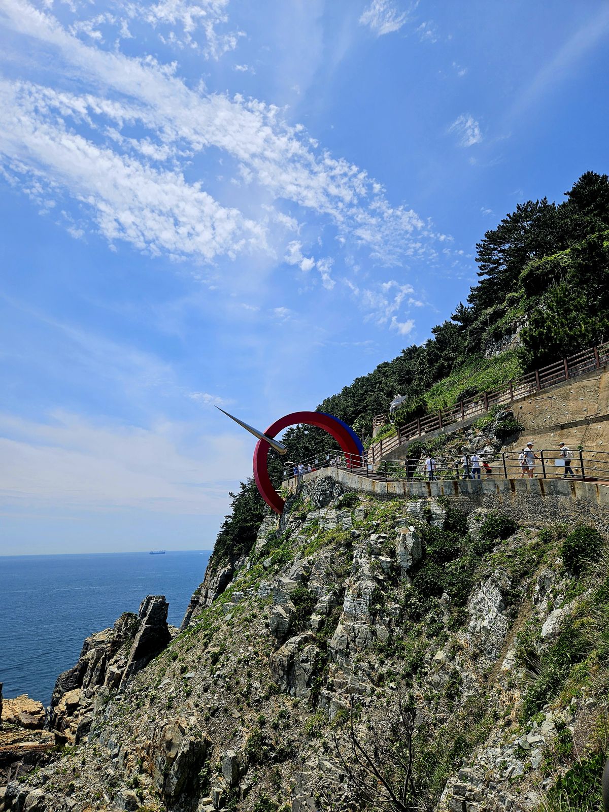 Un belvédère avec une sculpture rouge surplombant la mer, entouré d'une falaise rocheuse et d'une végétation verdoyante, sous un ciel bleu parsemé de nuages.