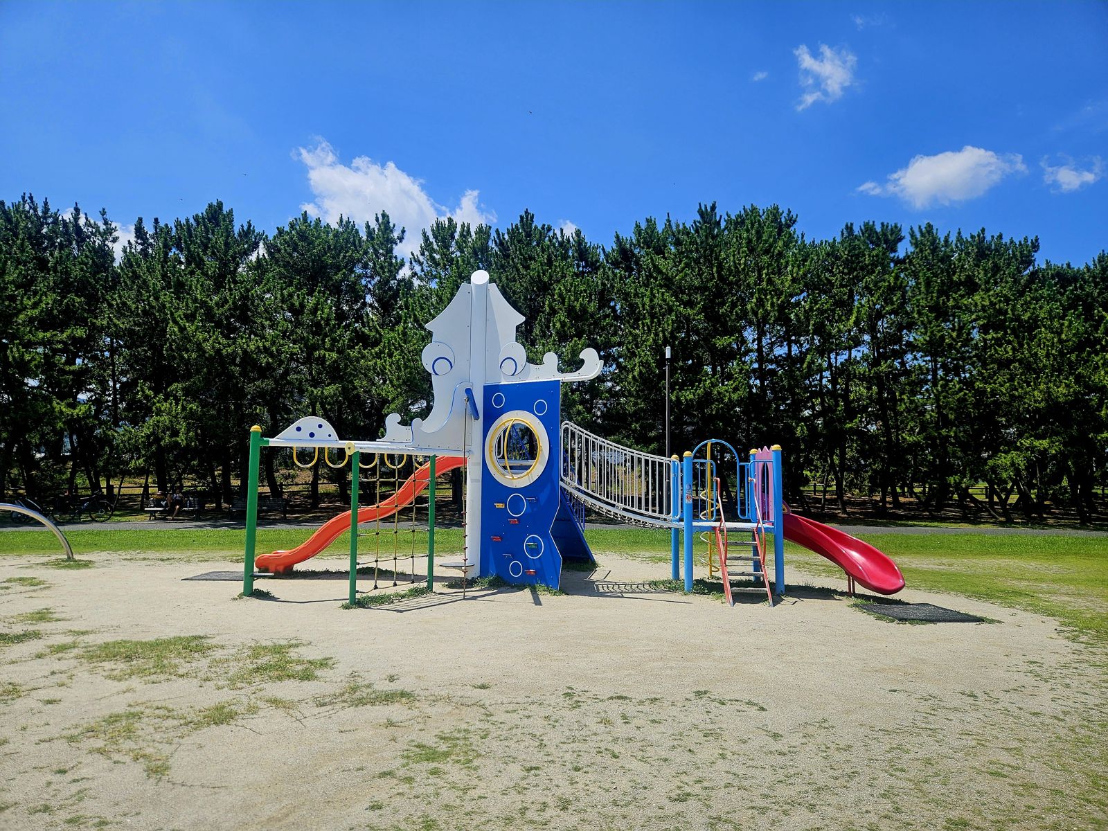 Aire de jeu colorée avec des toboggans et des balançoires, entourée d'arbres, sous un ciel bleu.