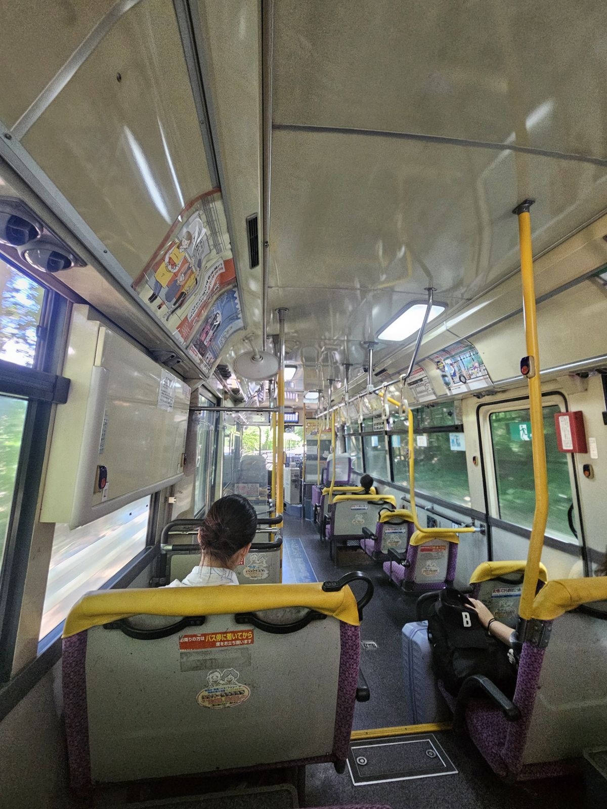 Intérieur d'un bus japonais avec des sièges occupés et vides, éclairé par la lumière naturelle. Des passagers sont assis, regardant à travers les fenêtres.