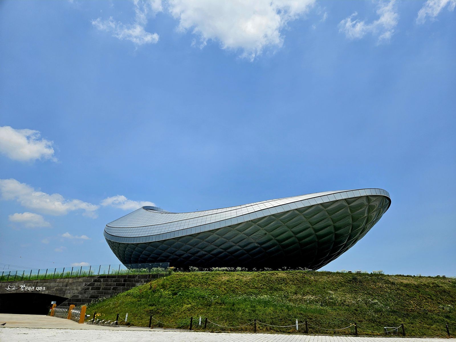 Vue d'un bâtiment moderne en forme de bateau, situé sur une colline verdoyante, sous un ciel bleu avec quelques nuages.