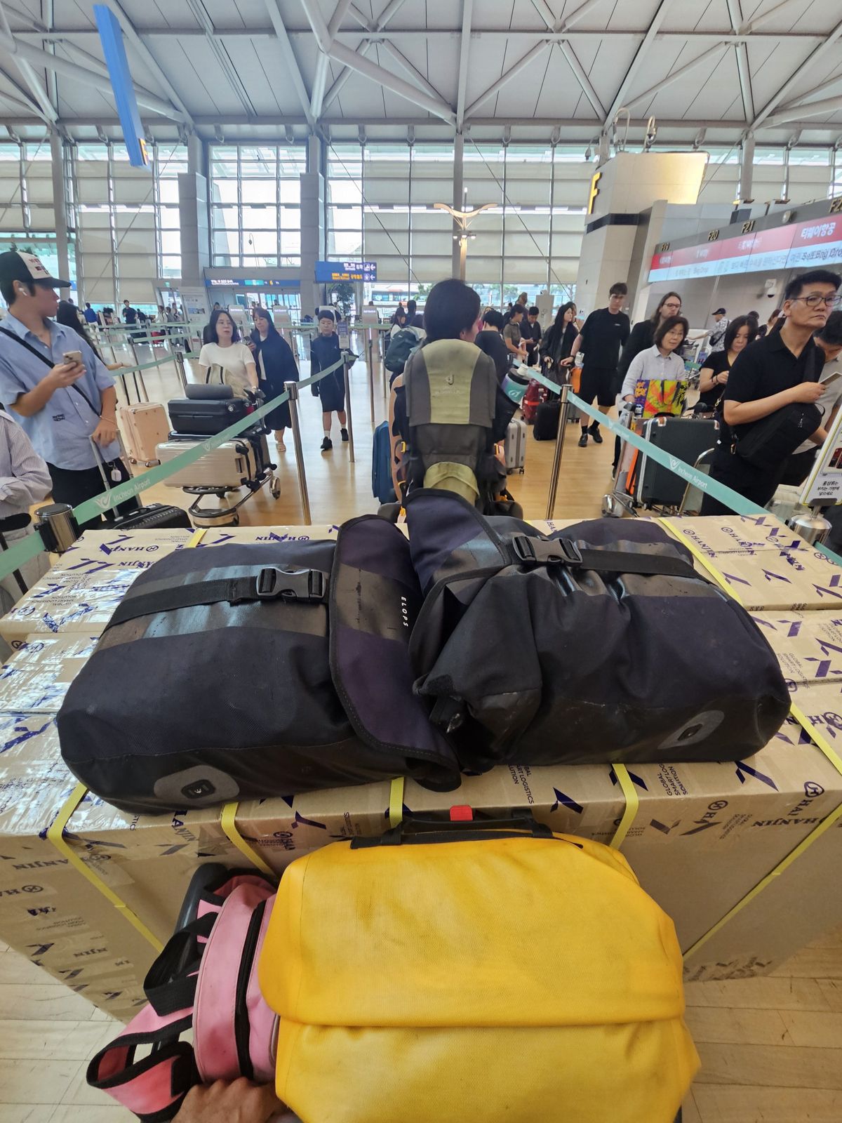 Photo d'un comptoir d'enregistrement à l'aéroport, avec des bagages empilés, dont des sacoches de vélo et un carton, dans une file d'attente.