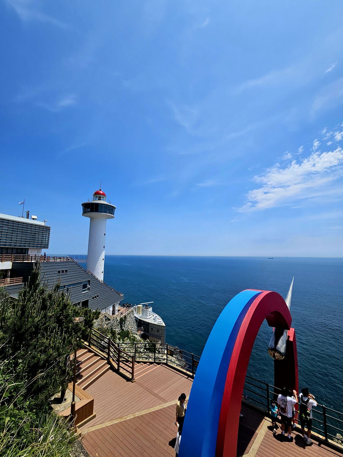 Vue panoramique d'un phare avec un ciel bleu clair et l'océan en arrière-plan, des touristes admirant le paysage près d'un arc coloré.