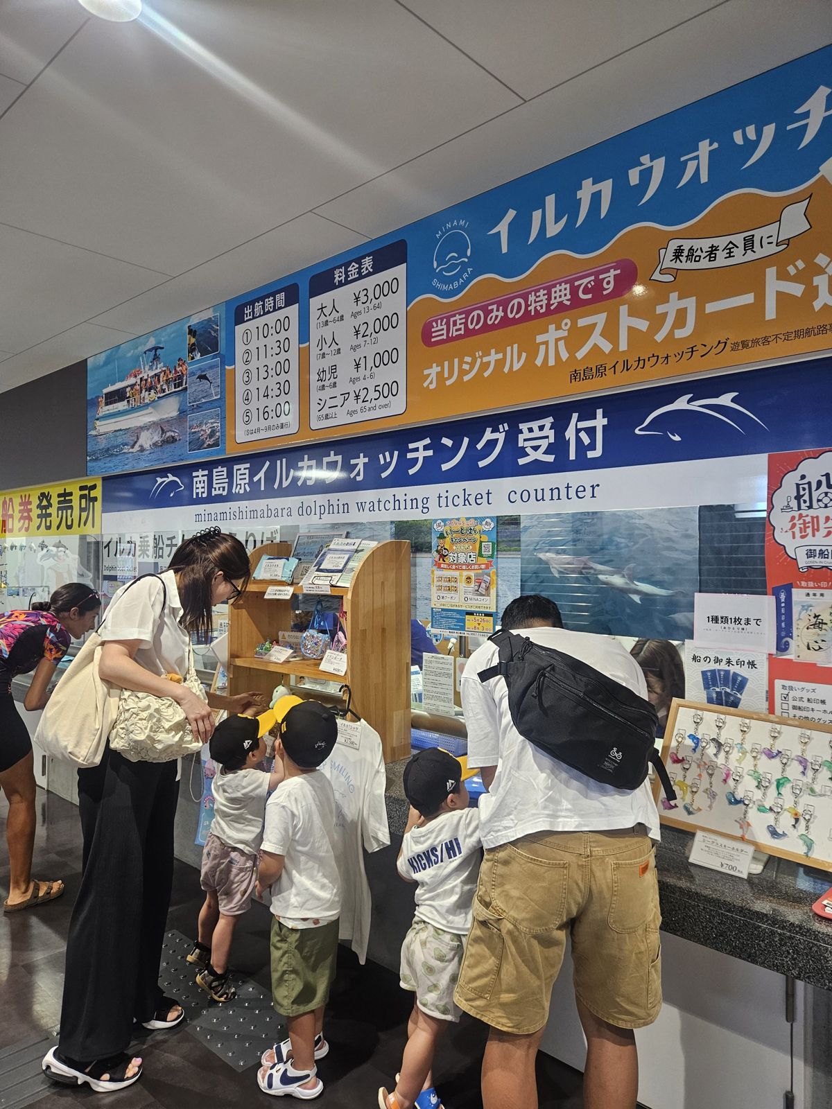Une famille se tient devant un comptoir de vente de billets pour des excursions d'observation de dauphins. On peut voir des panneaux d'affichage avec des horaires et des prix, ainsi que des brochures et des souvenirs. Les enfants, portant des casquettes, semblent intéressés par les informations affichées.