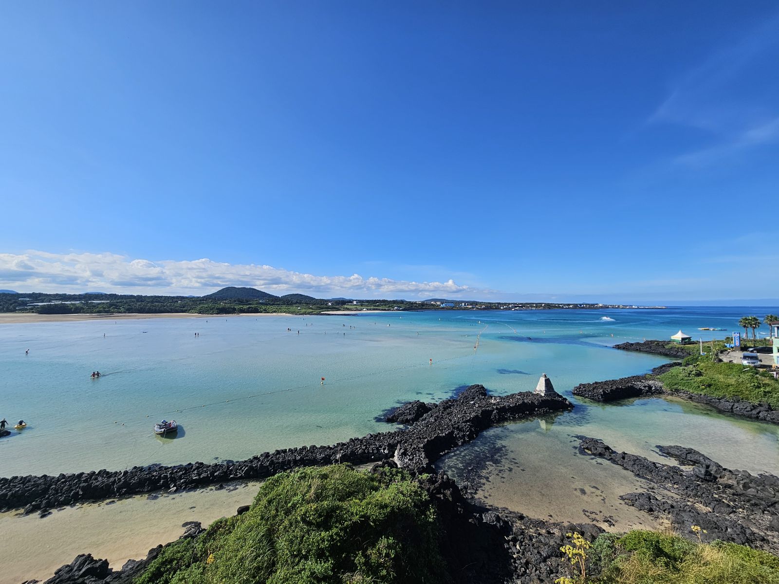 Vue panoramique de la plage de Sogeummak à Jeju, avec une mer claire et peu profonde, des formations rocheuses noires au premier plan et des personnes se baignant au loin sous un ciel bleu.
