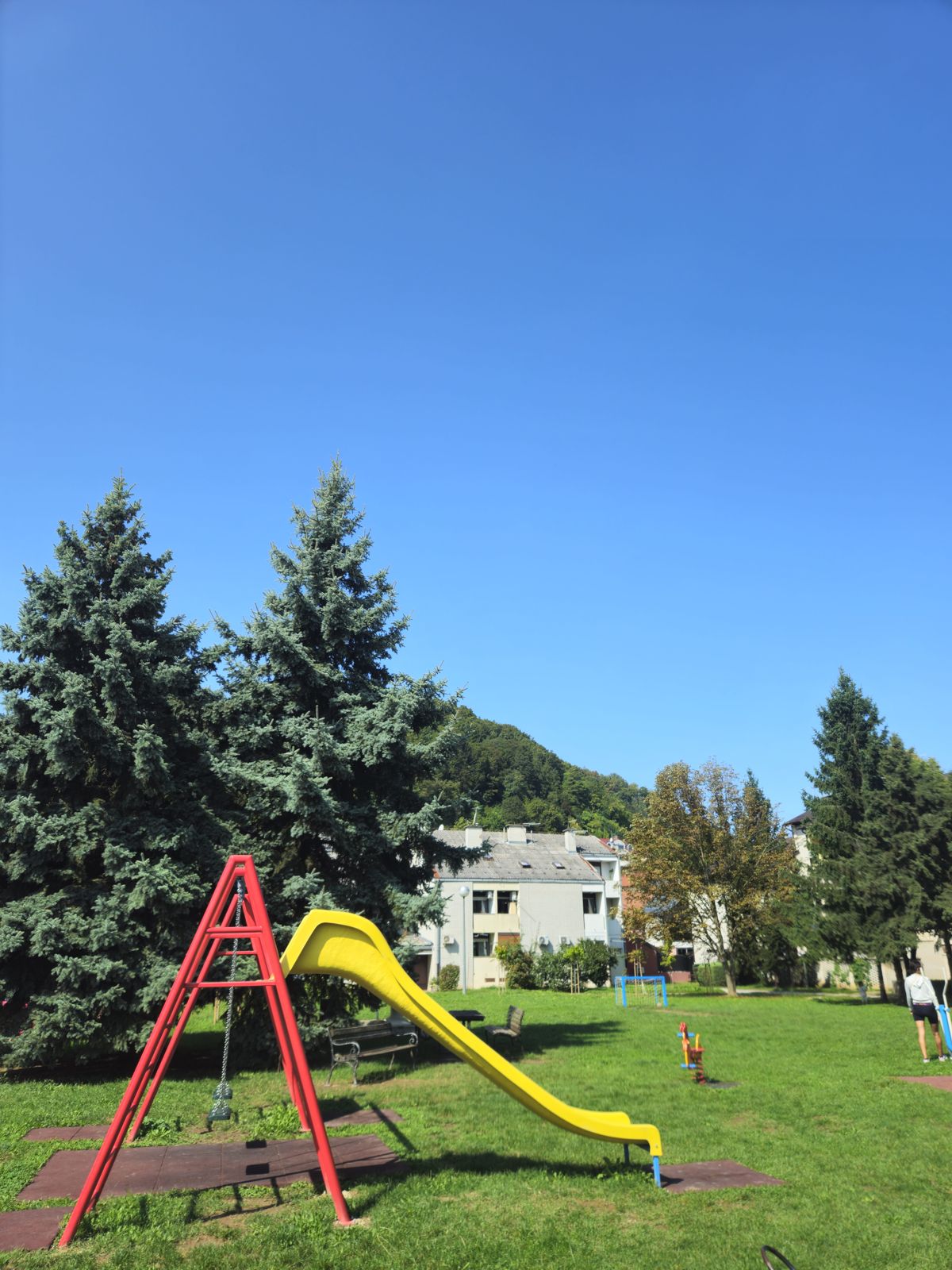 Une aire de jeux avec un toboggan jaune et une structure rouge, entourée d'arbres et d'un ciel bleu.