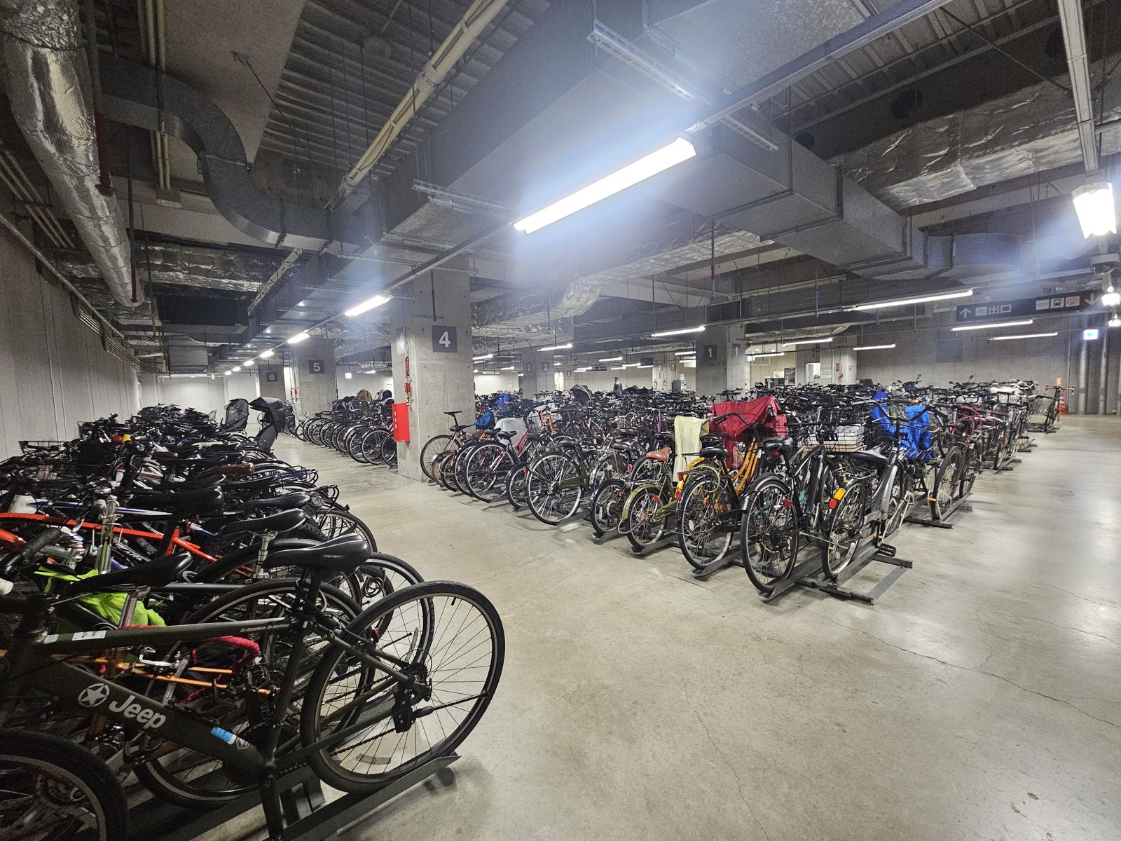 Un grand parking à vélos avec de nombreuses bicyclettes alignées, sous un éclairage fluorescent dans un espace urbain.