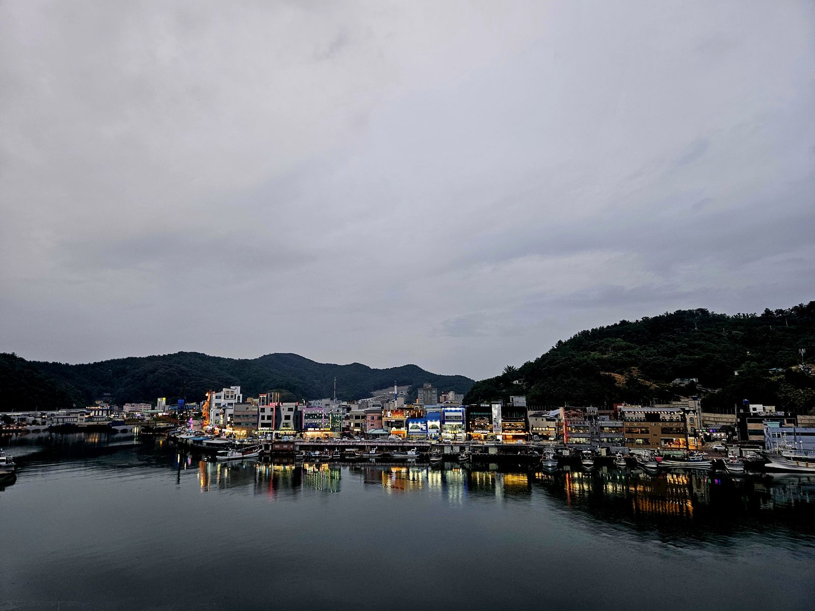 Vue panoramique d'une ville portuaire coréenne avec des bâtiments illuminés le long de la côte, reflétés dans l'eau calme sous un ciel nuageux.