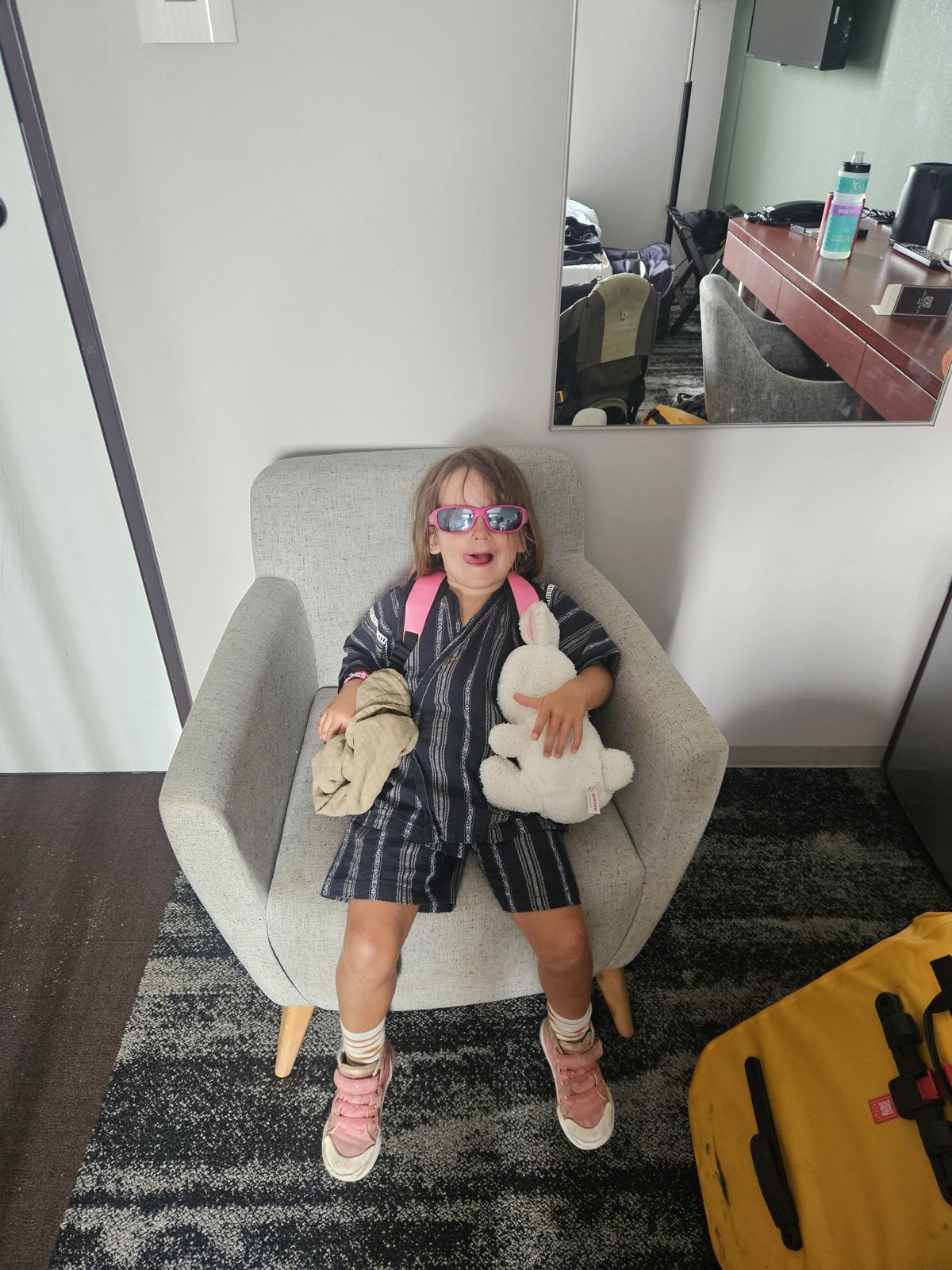 Une petite fille assise sur un fauteuil dans une chambre d'hôtel, portant des lunettes de soleil, un vêtement en tissu léger, et tenant un doudou et un vêtement plié.