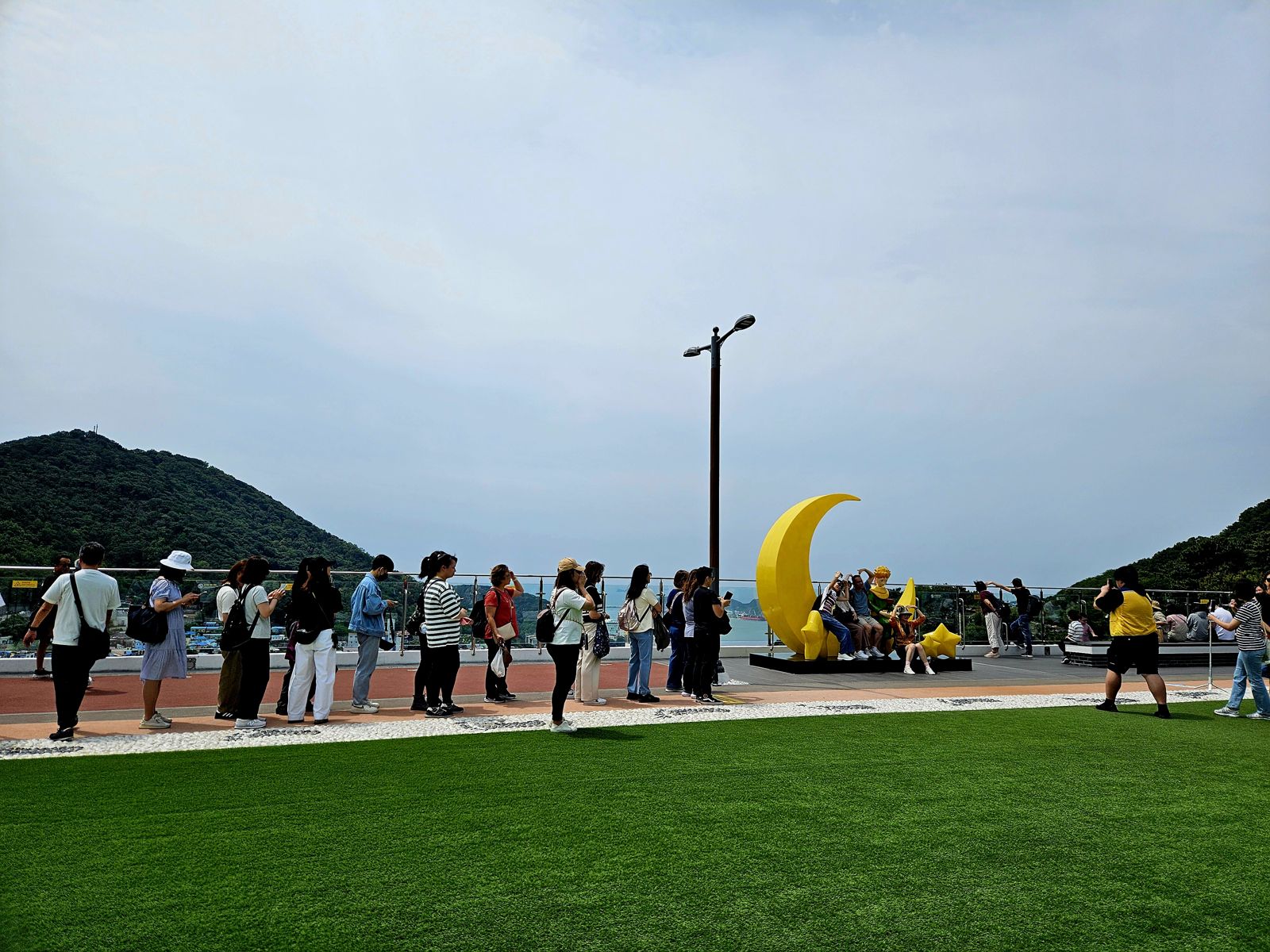 Un groupe de personnes fait la queue pour prendre des photos avec des statues inspirées du Petit Prince, au bord de la mer, avec des collines en arrière-plan.