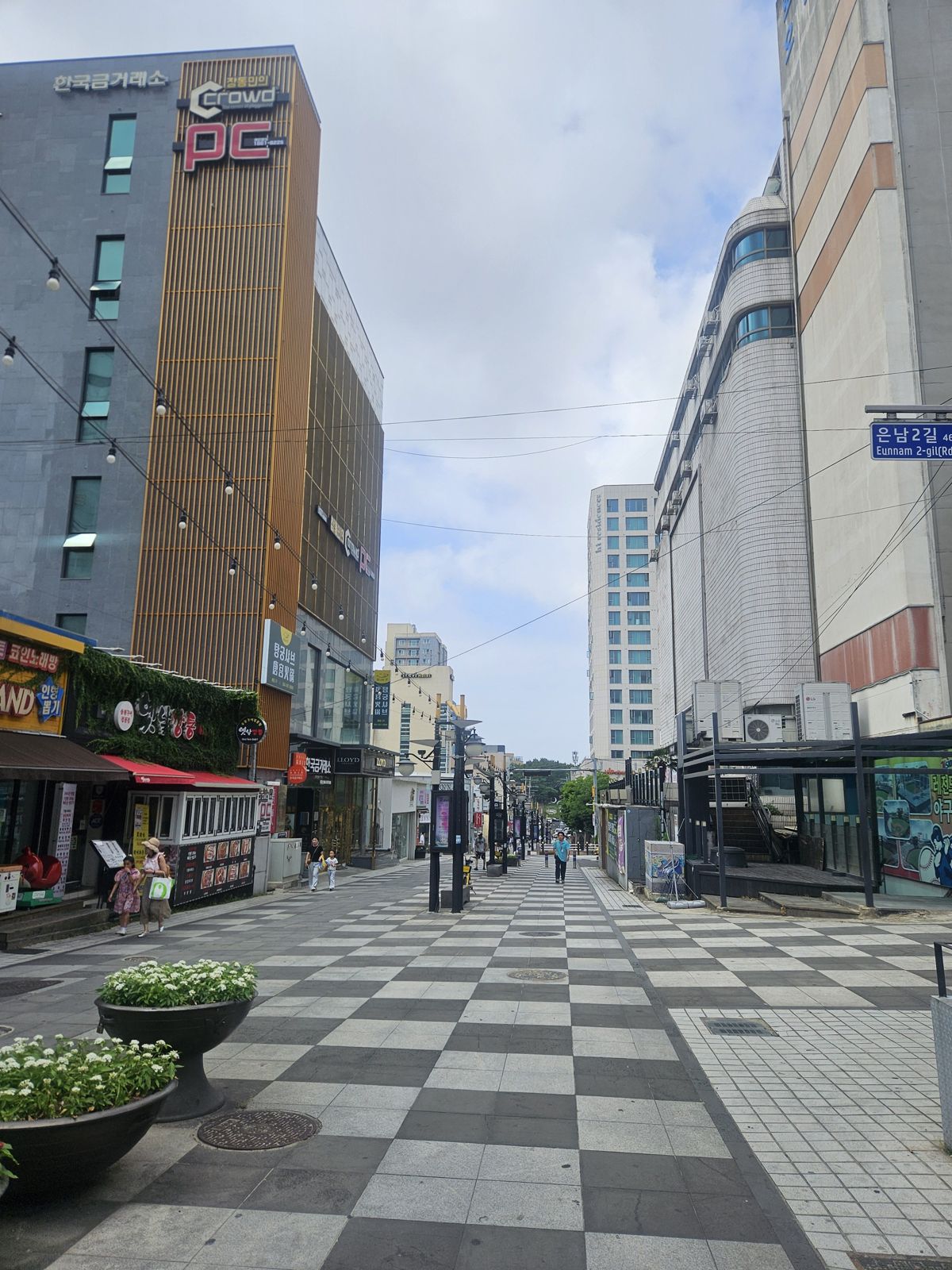 Vue d'une rue moderne et peu fréquentée à Jeju, avec des bâtiments commerciaux et des espaces verts, sous un ciel partiellement nuageux.