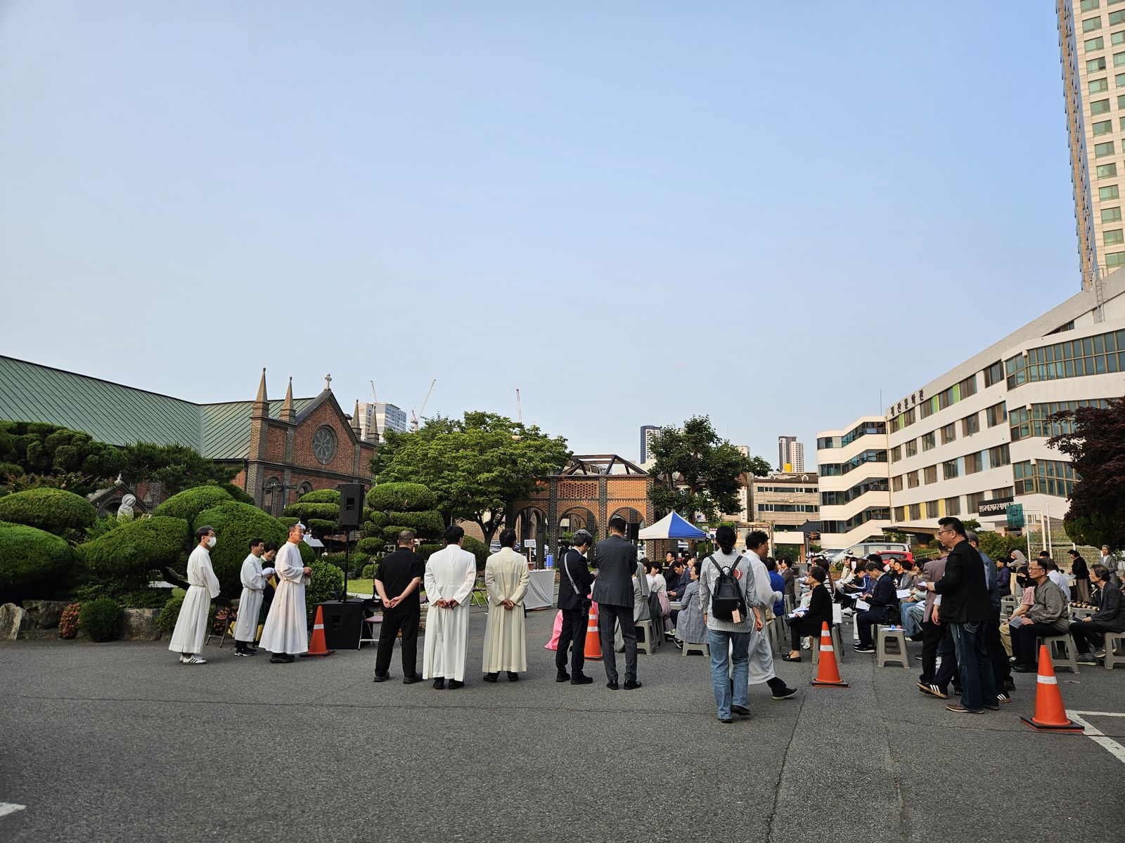 Un groupe de personnes assiste à un mariage à l'extérieur d'une église, avec des chaises disposées en cercle et des cônes de signalisation autour. Quelques clergés en tenue cérémonielle sont présents, et des bâtiments modernes se trouvent en arrière-plan.