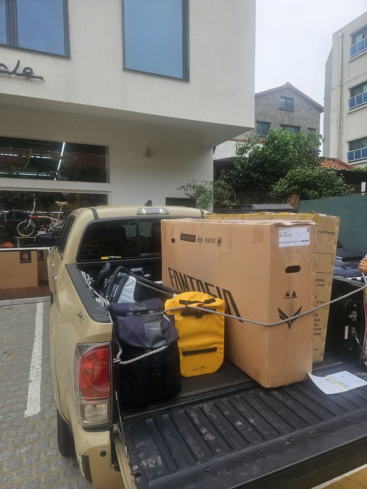 Un pick-up stationné devant un magasin de vélos avec des cartons de vélos attachés à l'arrière, accompagnés de quelques sacs de transport.