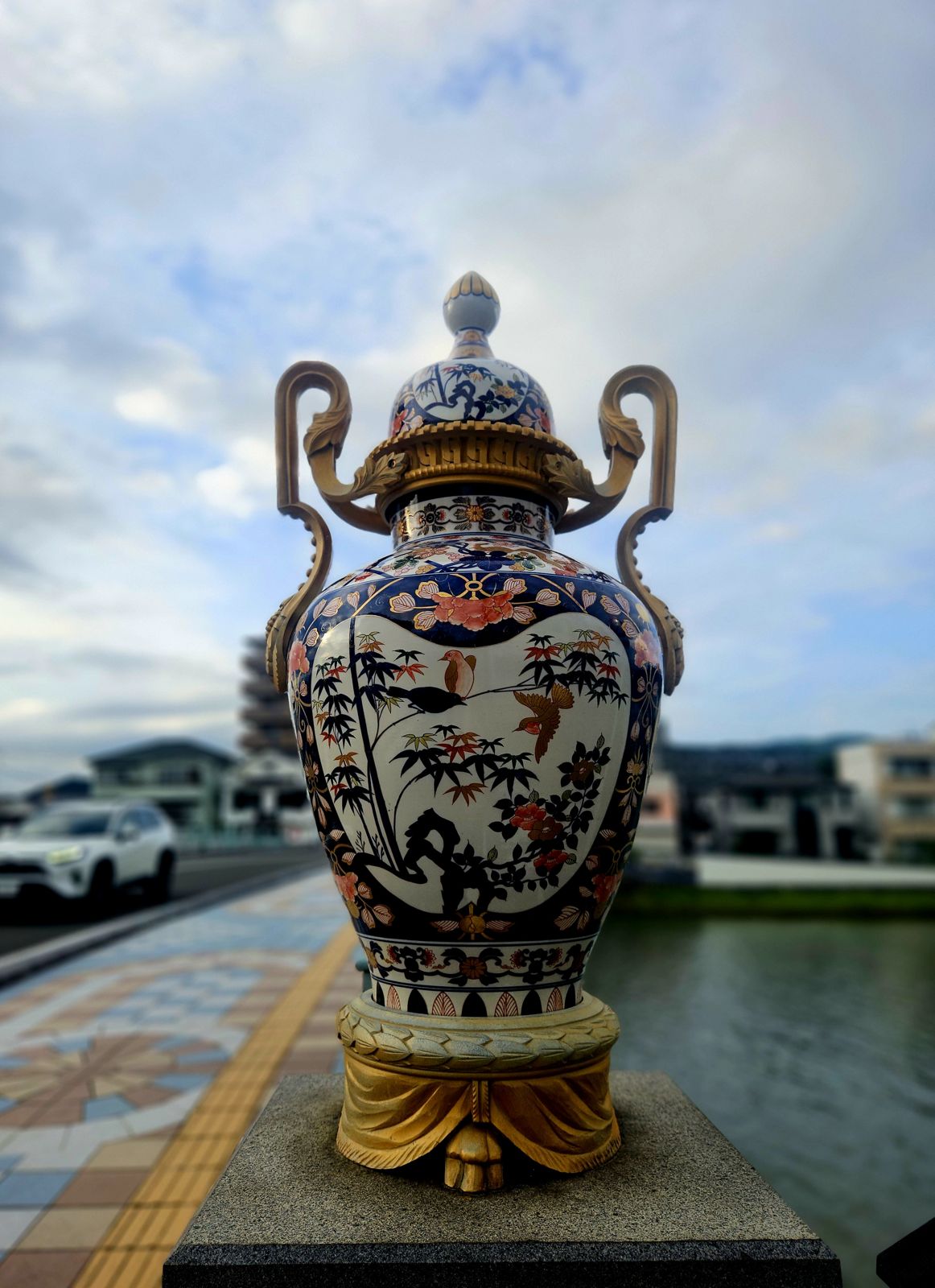 Vase en porcelaine décoratif, vue avant, situé sur un trottoir près d'un canal avec un ciel nuageux en arrière-plan.