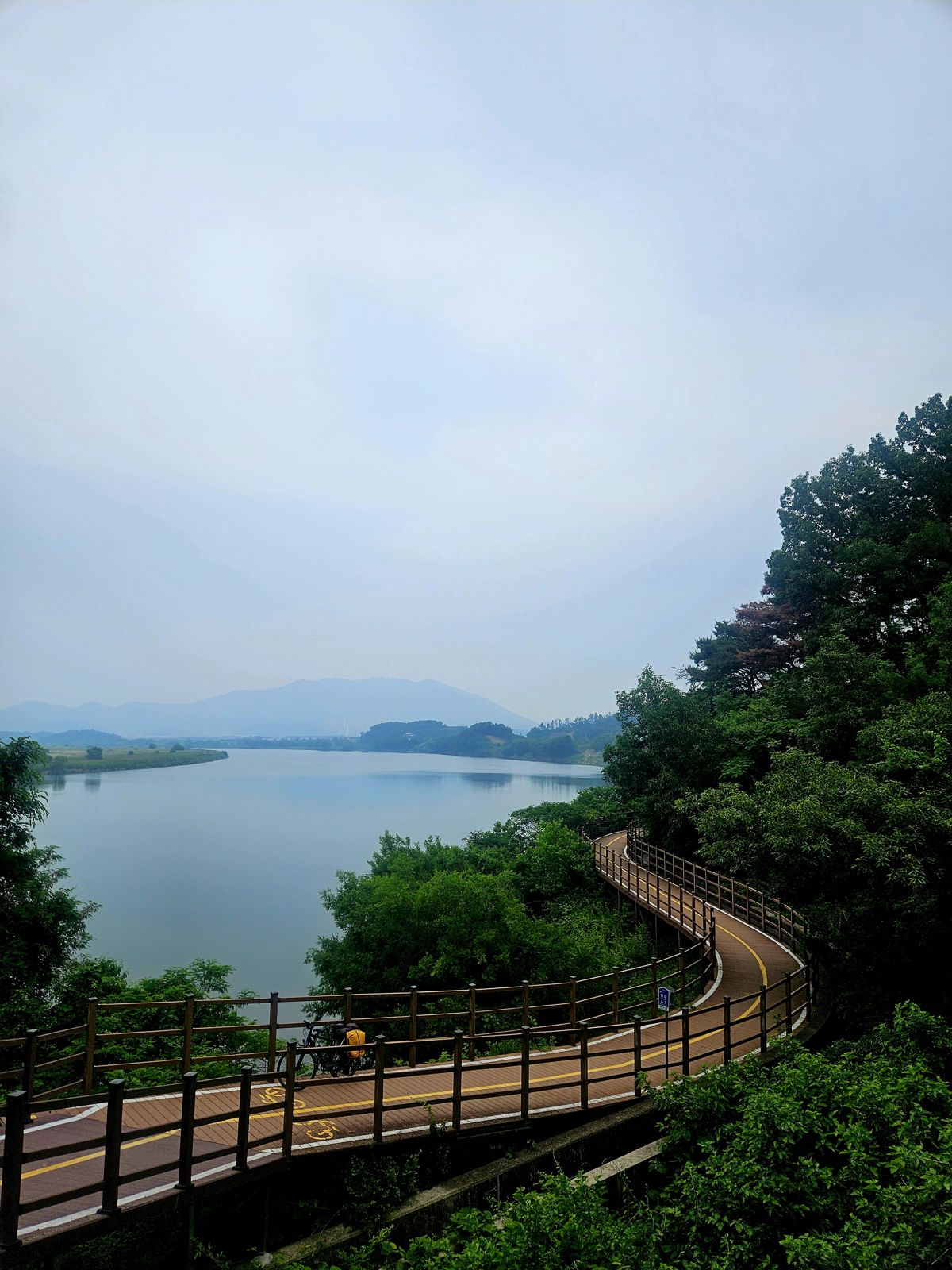 Vue panoramique d'une véloroute serpentant le long d'un plan d'eau, entourée de verdure et de collines, avec un ciel nuageux.