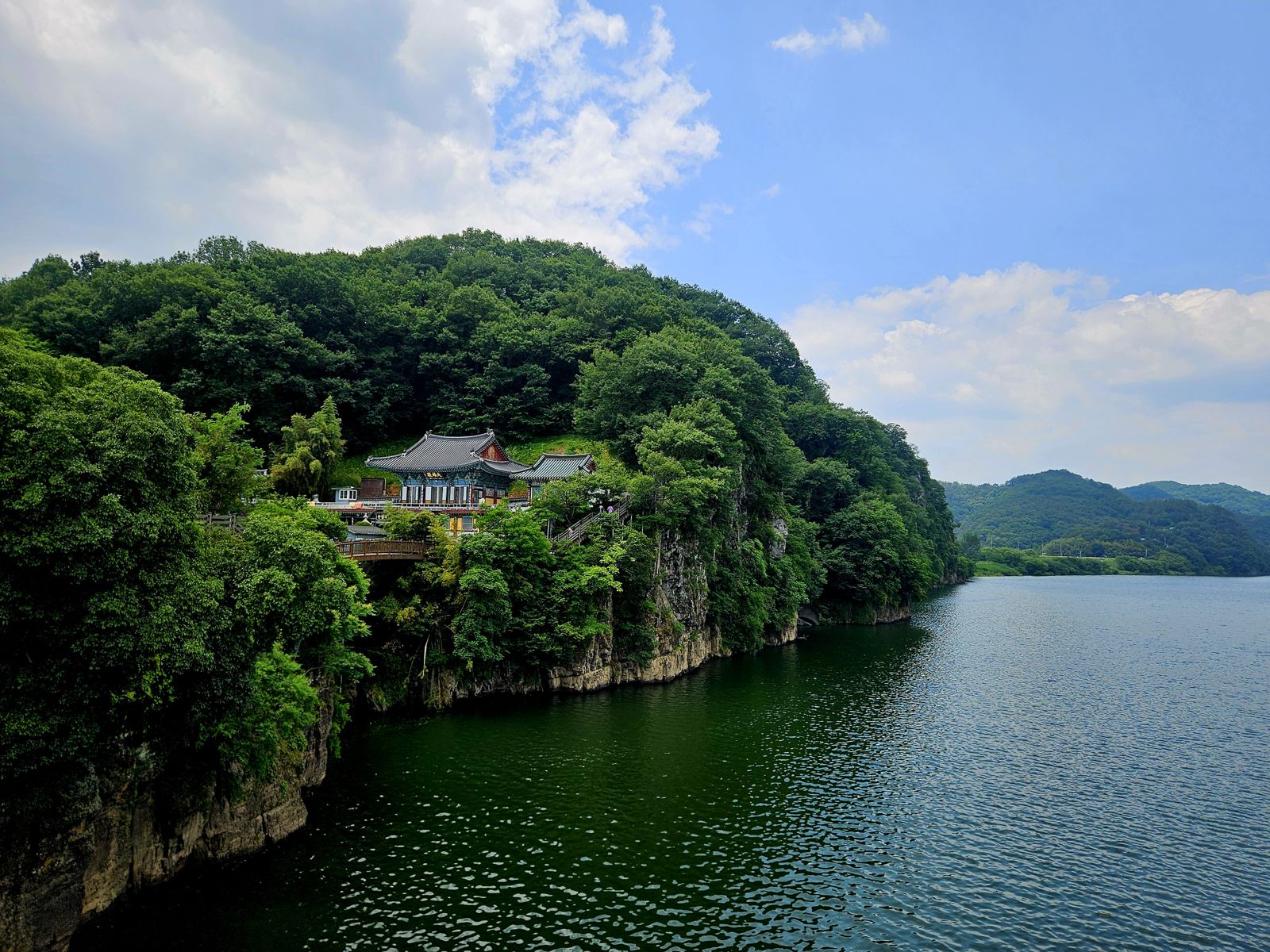 Vue d'une maison traditionnelle coréenne perchée sur une falaise verdoyante surplombant un lac calme sous un ciel nuageux.